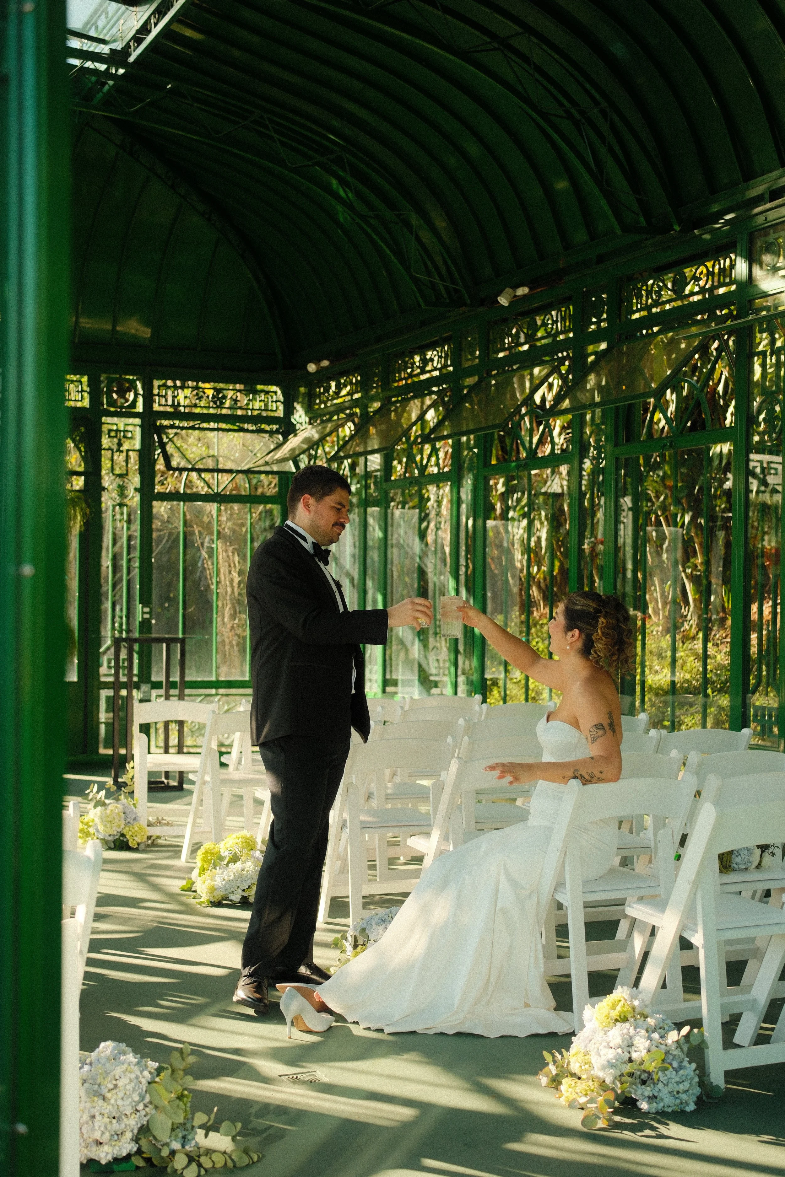 Novios compartiendo un momento durante su ceremonia de boda en un espacio tipo invernadero, rodeados de flores y luz natural.
