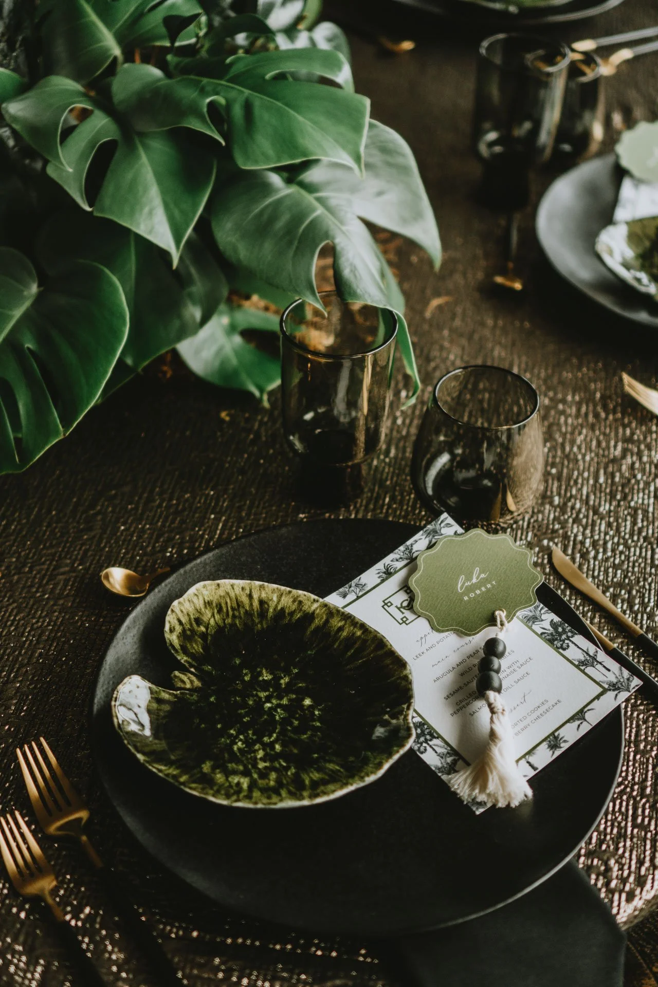 A modern table setting with a large green monstera plant in the background, black and gold flatware, a black plate with a green and black ceramic bowl, black-tinted glasses, a printed menu with a green name tag labeled 'Luka', and a decorative beaded tassel.