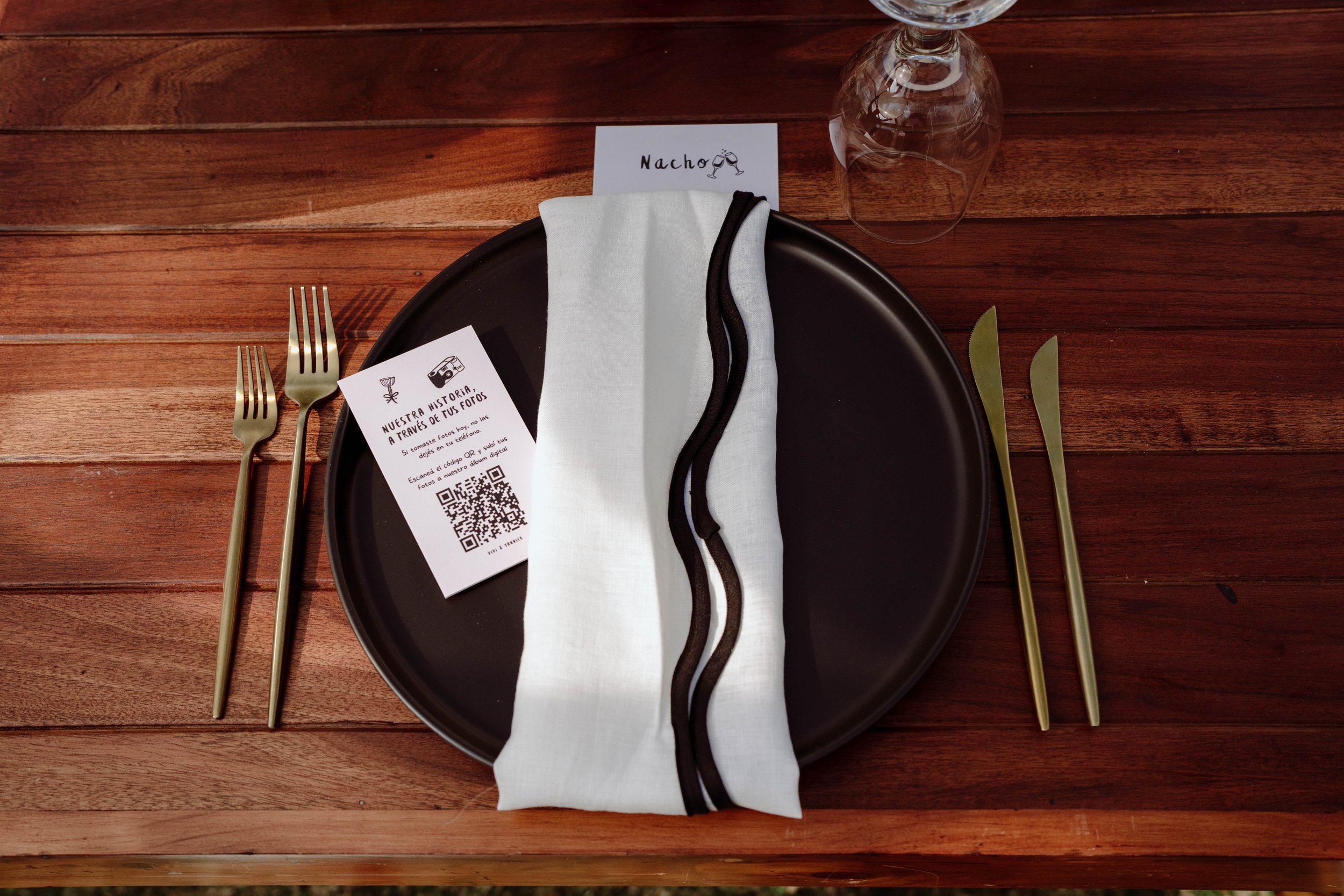 Detalle de puesto de mesa en una boda en Costa Rica, con plato negro, cubiertos dorados, tarjeta personalizada y código QR para compartir fotos.