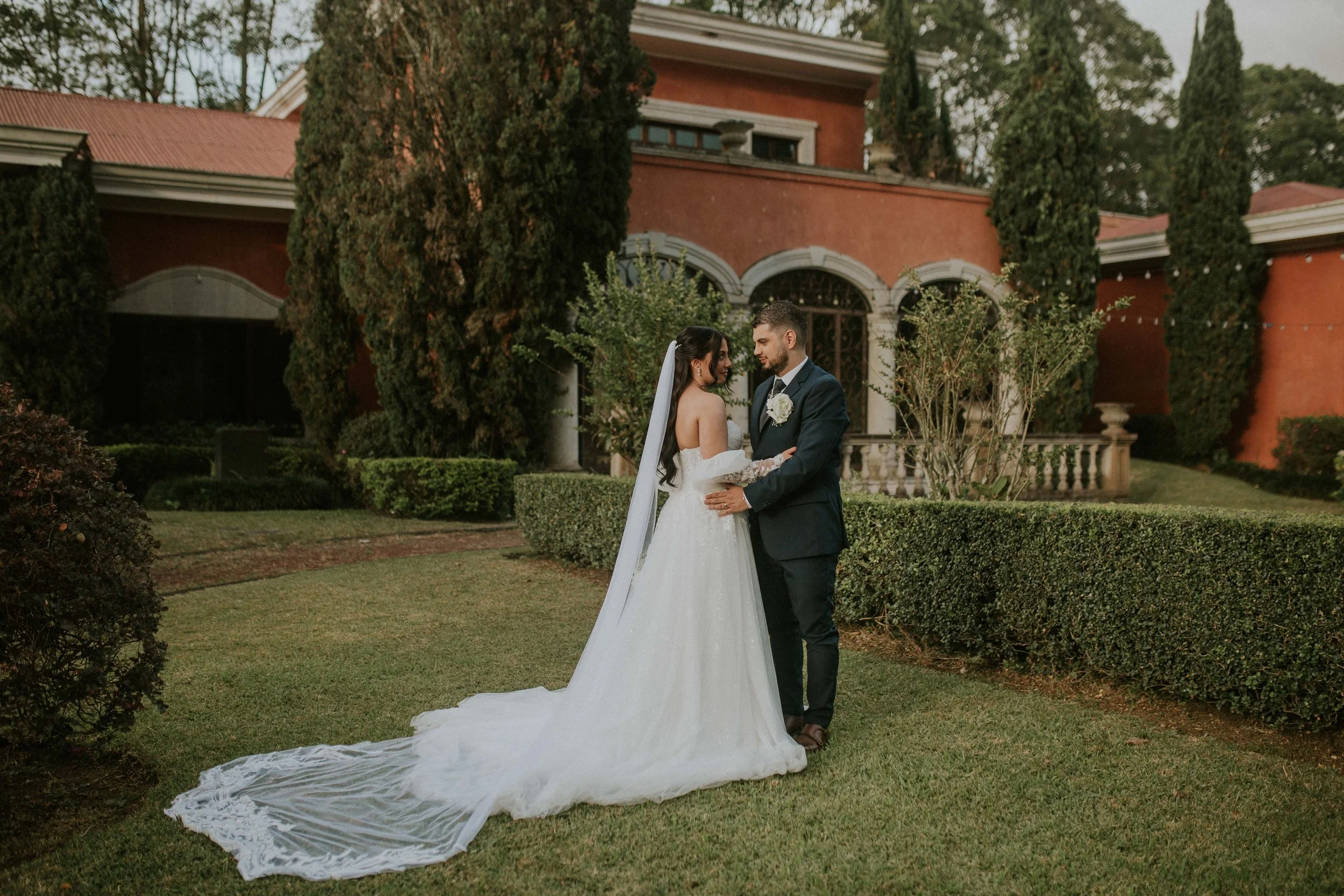 A bride and groom standing close together in a garden, gazing into each other's eyes during their wedding. The bride is wearing a white wedding gown with lace sleeves and a veil, while the groom is dressed in a dark suit with a boutonniere. Behind them is a large, elegant house with arched windows, surrounded by tall trees and lush greenery.