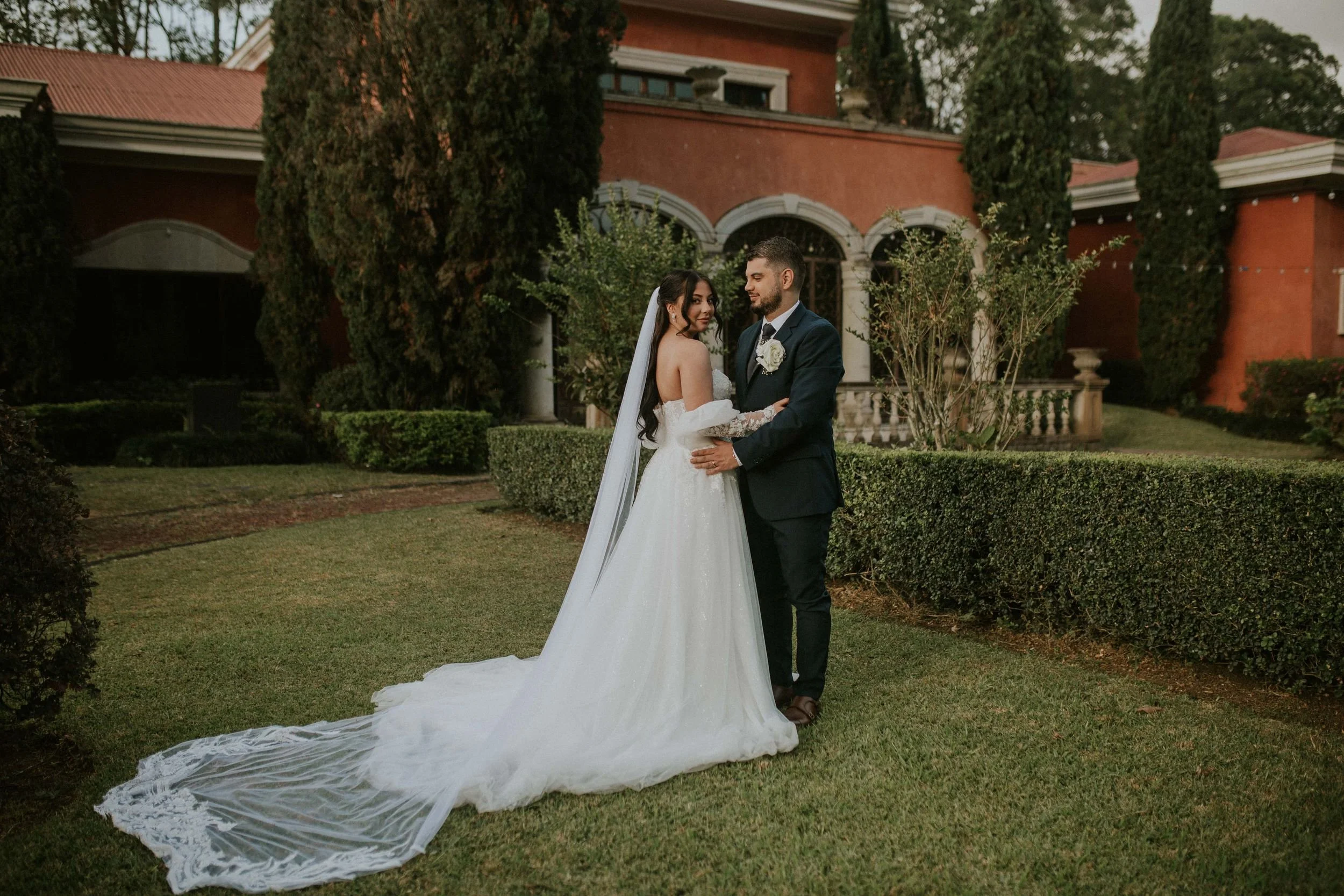 A bride and groom standing hand in hand on a lawn in front of a large red building with arched windows and tall trees, during a wedding celebration.