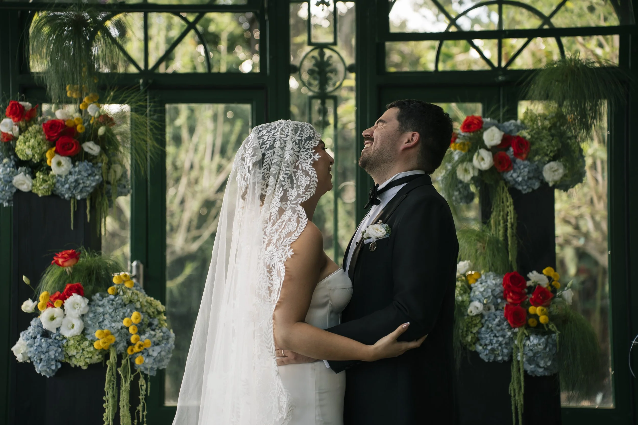Novios sonriéndose durante su ceremonia de boda, rodeados de arreglos florales coloridos en un invernadero lleno de luz natural.