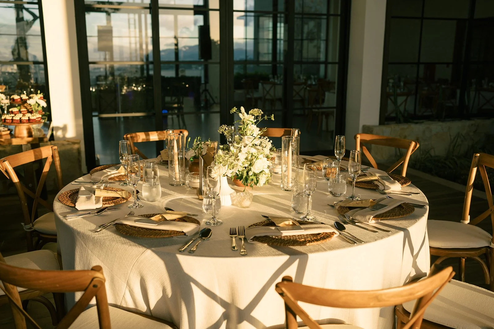 Montaje de mesa para recepción de boda en un espacio interior con ventanales, luz cálida, flores blancas y detalles elegantes.