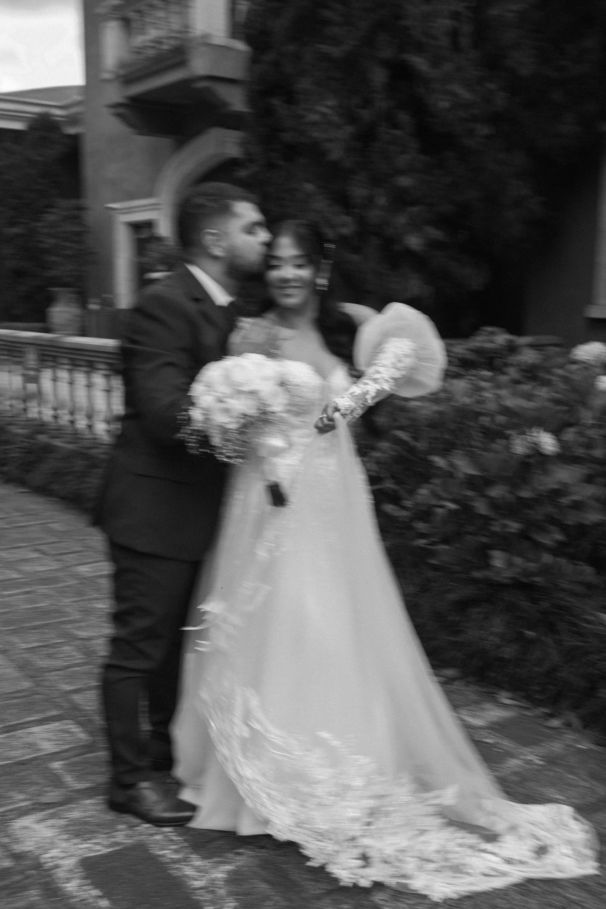 A black and white photo of a bride and groom on their wedding day. The groom is in a suit, holding a bouquet, and the bride is wearing a long wedding dress and holding a bouquet. They are standing outside near a house with a garden.