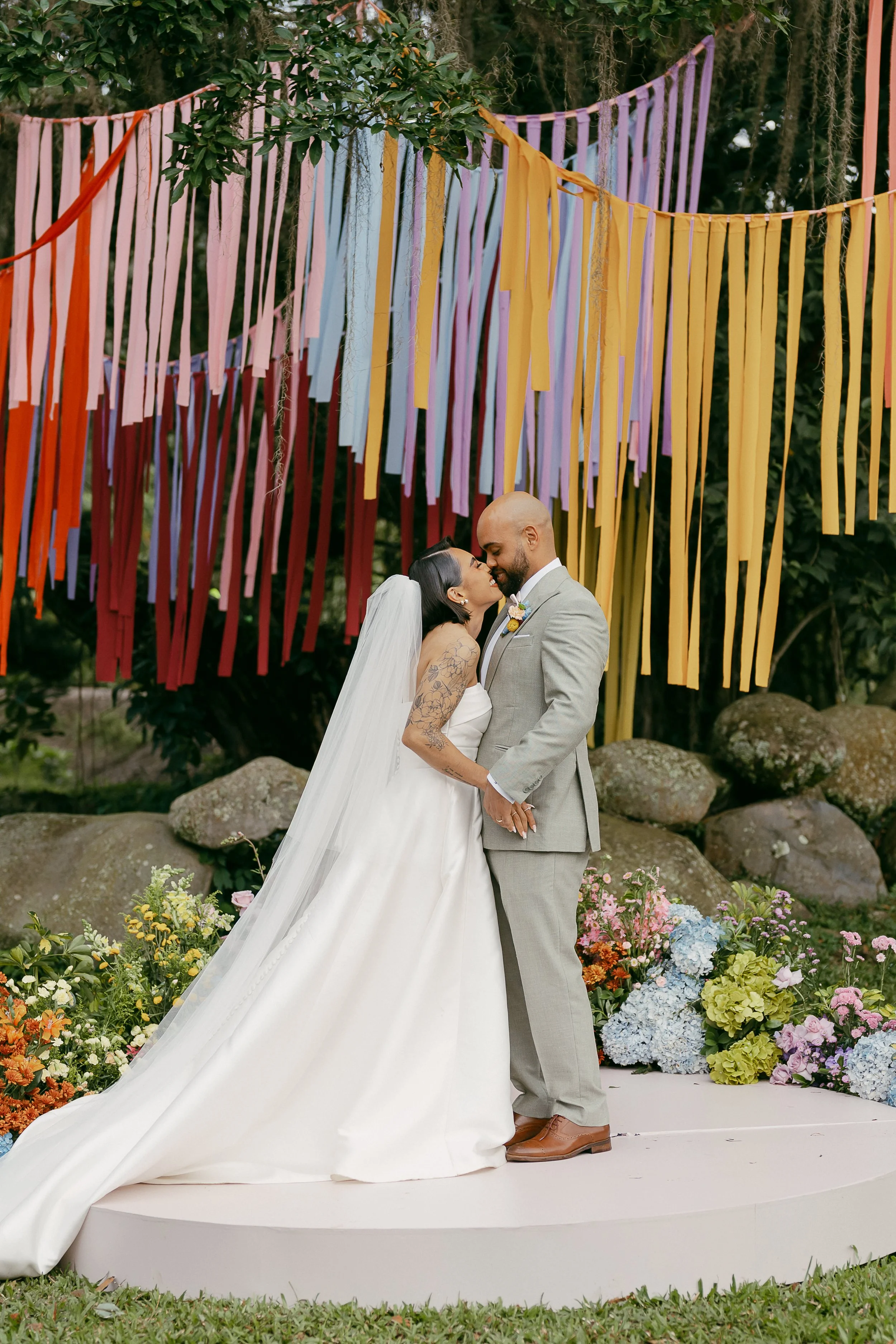 A bride and groom kissing during their wedding ceremony outdoors, standing on a white platform decorated with colorful flowers, with multicolored ribbons hanging overhead.