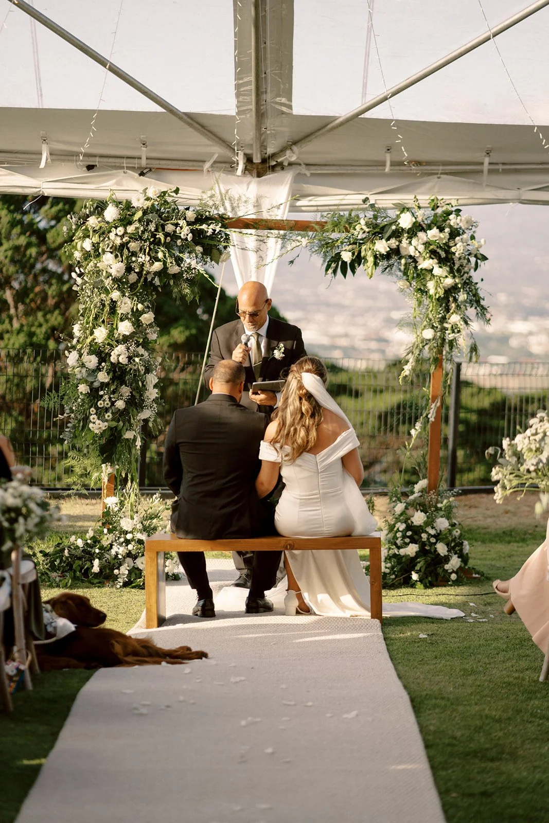 Ceremonia de boda al aire libre con los novios sentados frente al altar floral, acompañados por su perro en un momento lleno de calma y cercanía.
