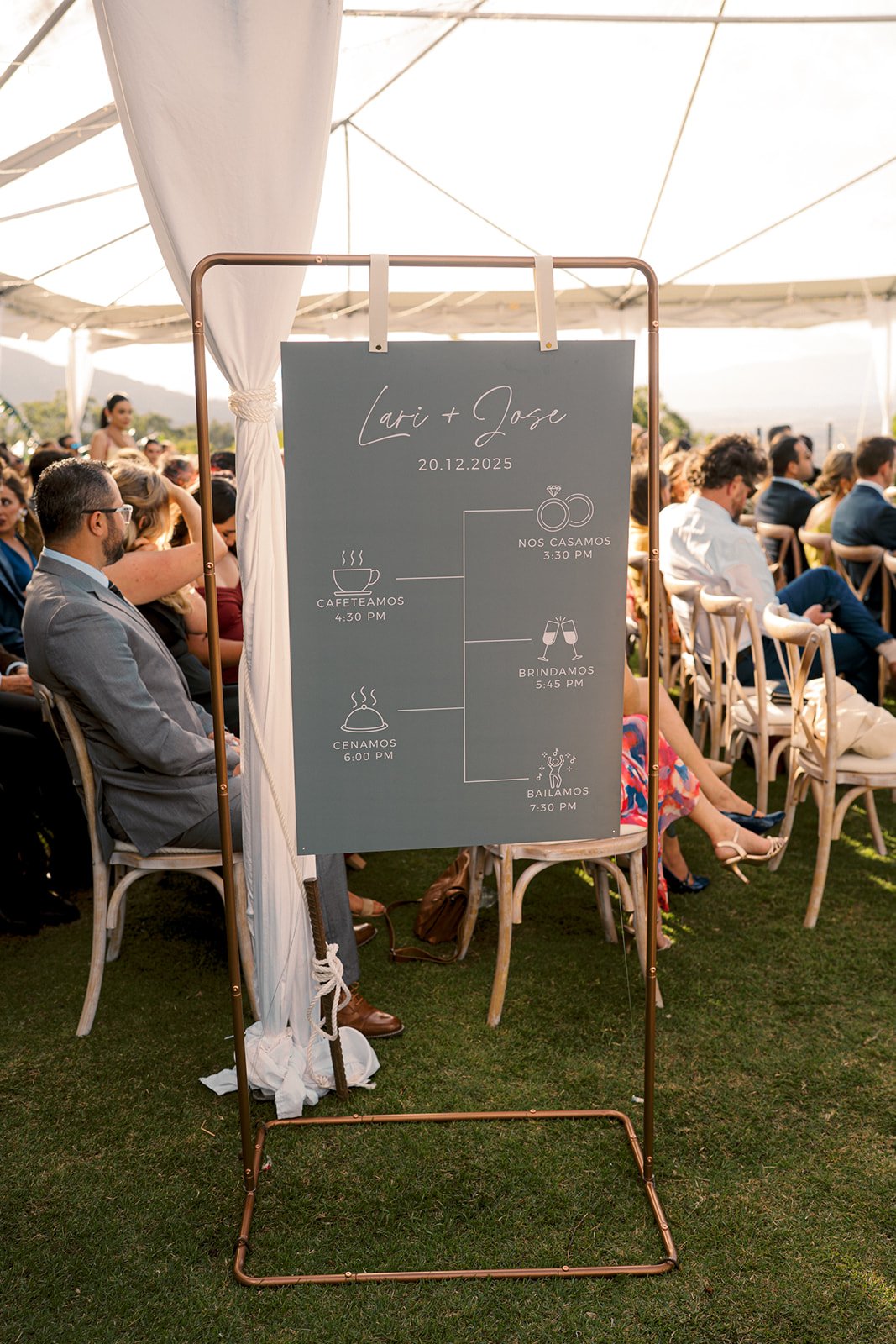 Letrero con el cronograma de la recepción durante una boda al aire libre, con mesas e invitados bajo una carpa blanca.