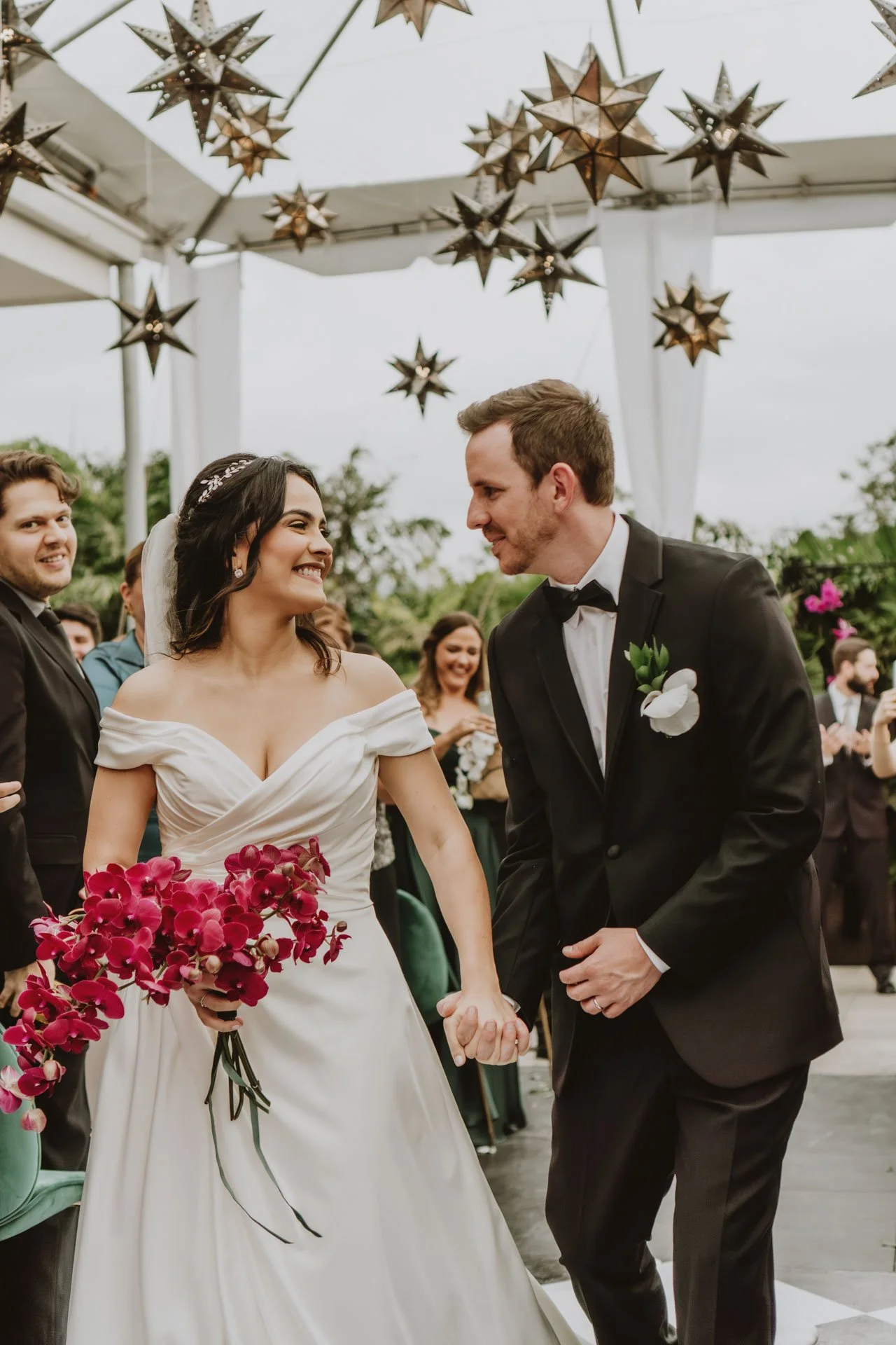 Pareja sonriendo durante su ceremonia de boda, con bouquet rosado, invitados al fondo y lámparas decorativas en forma de estrella.