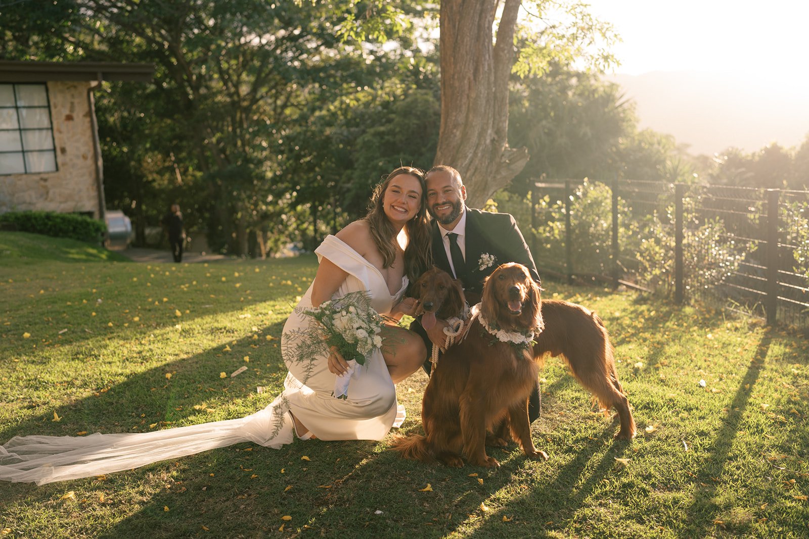 Novios arrodillados junto a sus dos perros durante una sesión al aire libre al atardecer, en un momento lleno de alegría y cercanía.