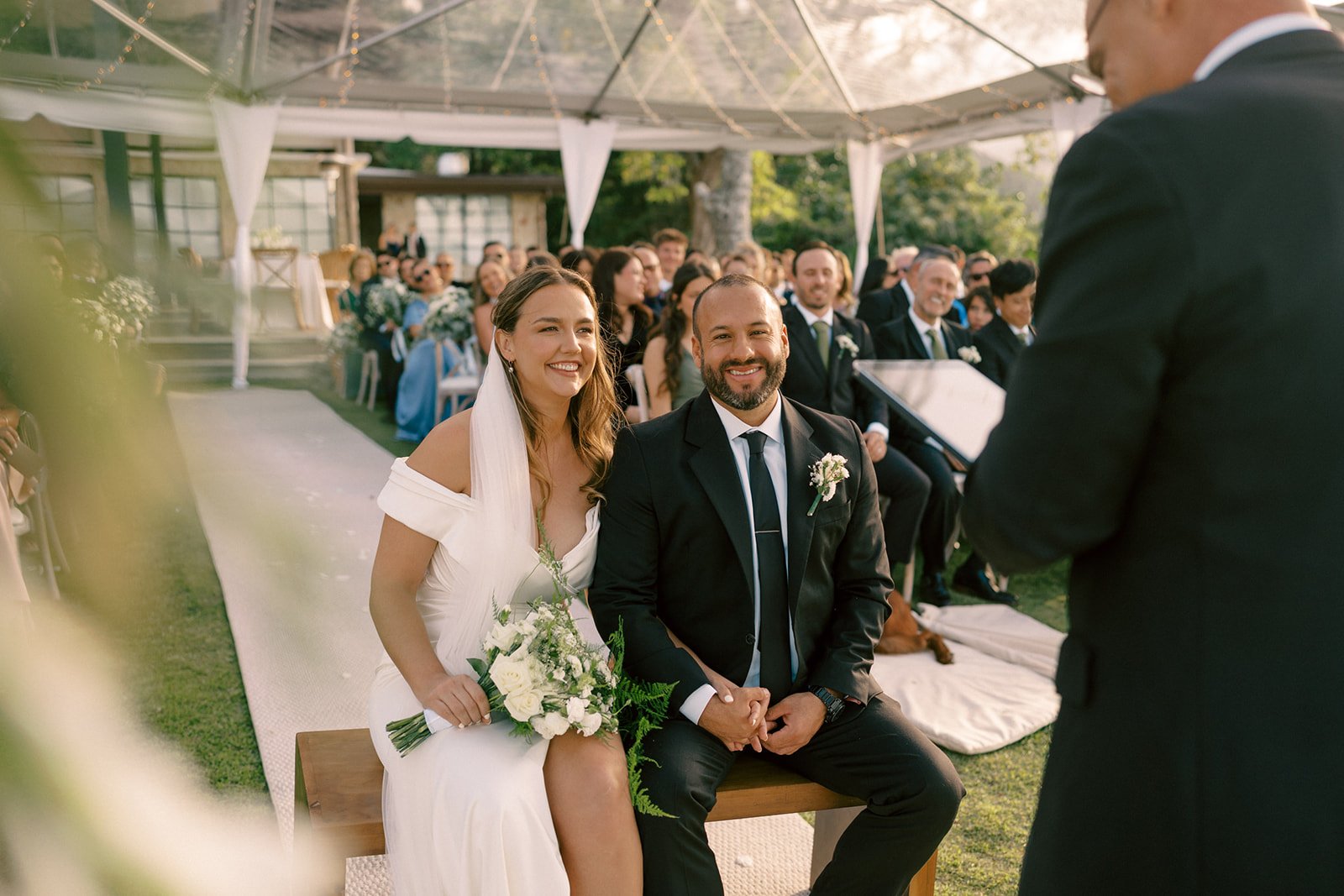 Pareja sentada durante la ceremonia de su boda, con la novia sosteniendo su bouquet y los invitados observando al fondo bajo una carpa.