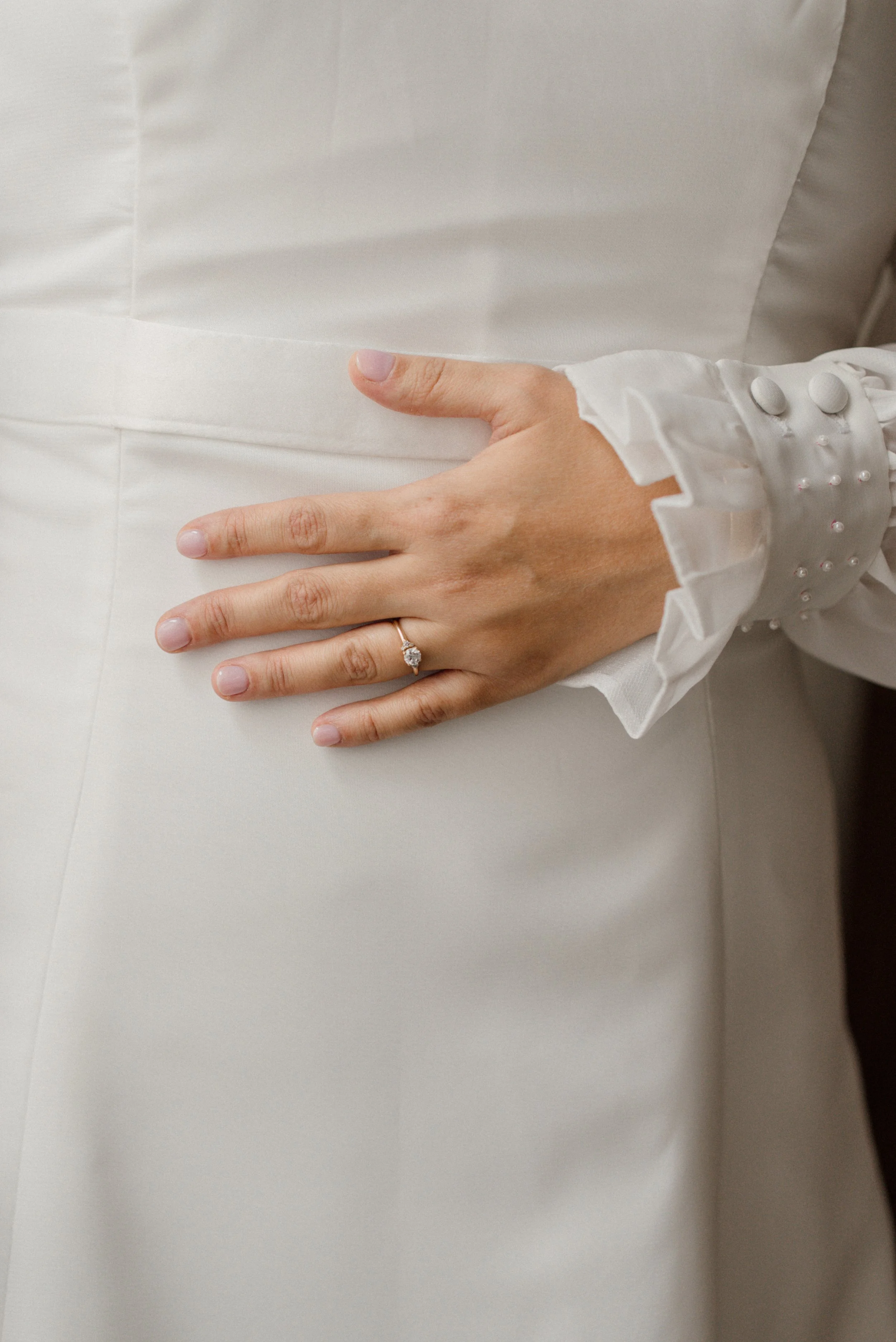 Close-up of a woman's hand with an engagement ring resting on the waist of a white dress, showing a white blouse with pearl button details and ruffled cuffs.