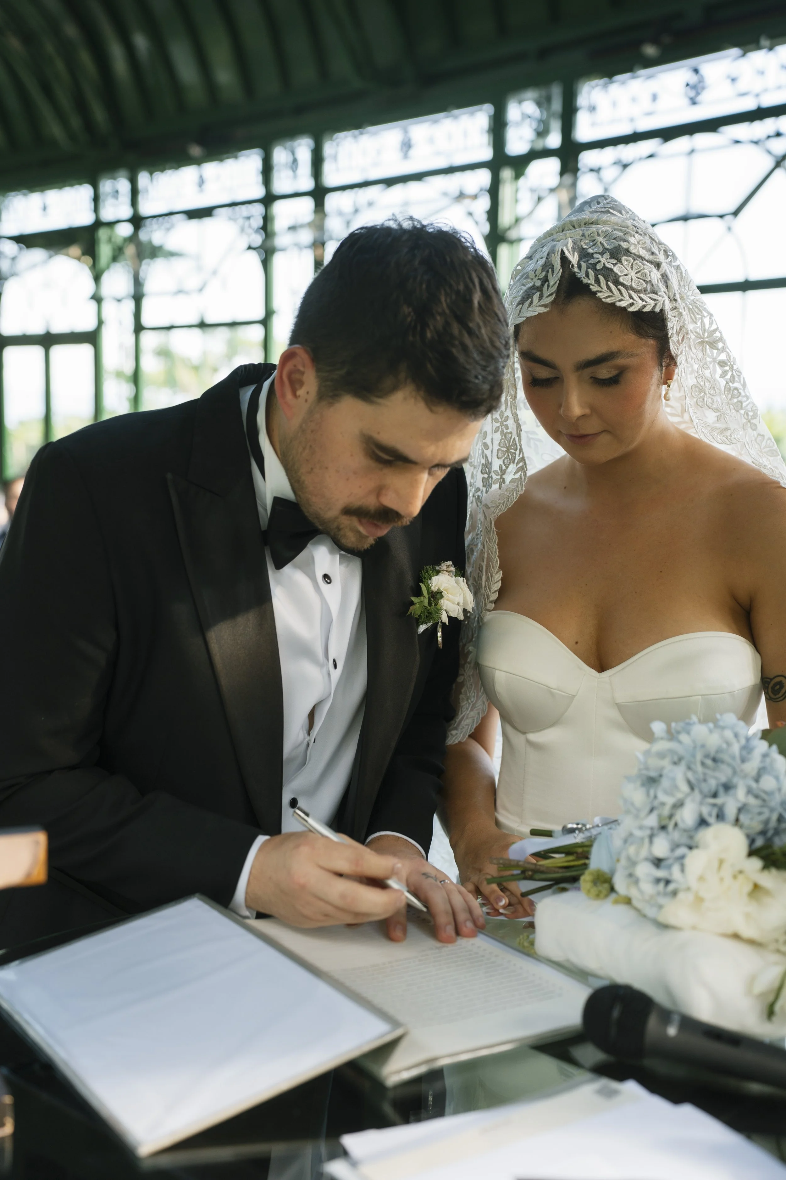 Firma de documentos durante la ceremonia de boda, con la novia acompañando al novio en un espacio interior con grandes ventanales.