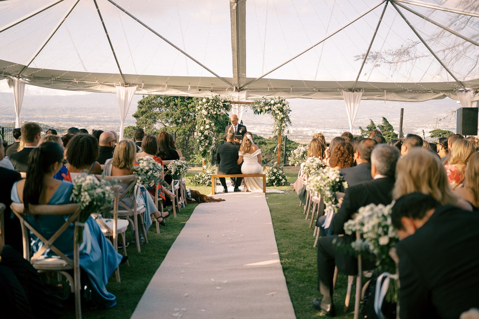 Ceremonia de boda bajo carpa, con los novios en el altar, invitados sentados a ambos lados y un arco floral como fondo.
