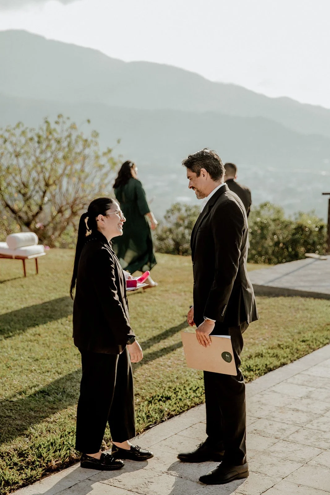 A man and a woman in business attire standing outdoors, smiling, and talking to each other on a sunny day with mountains in the background.