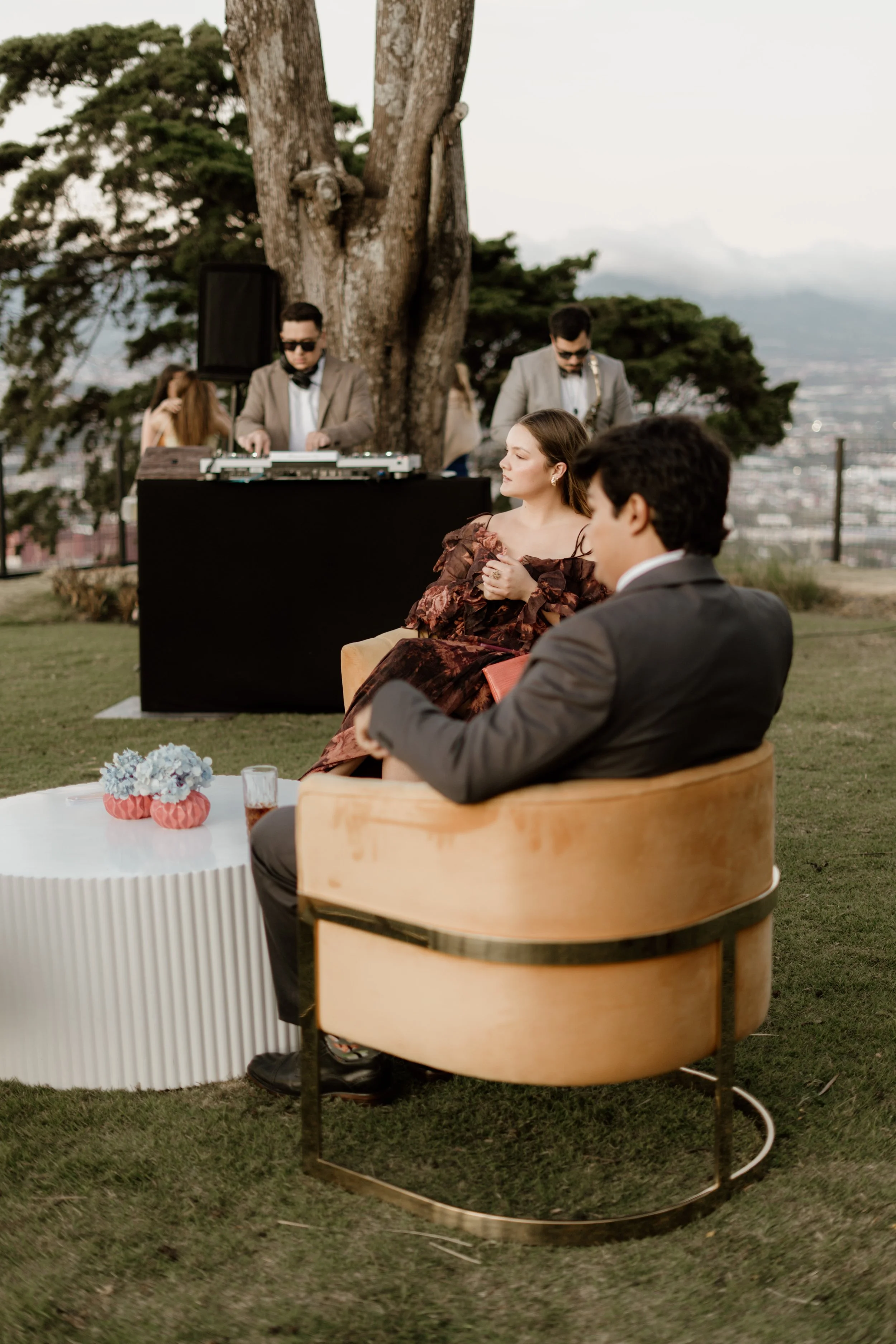 Couple sitting on a tan velvet chair and a cream-colored side table with flowers, enjoying an outdoor gathering. DJ and other guests are in the background under a large tree.
