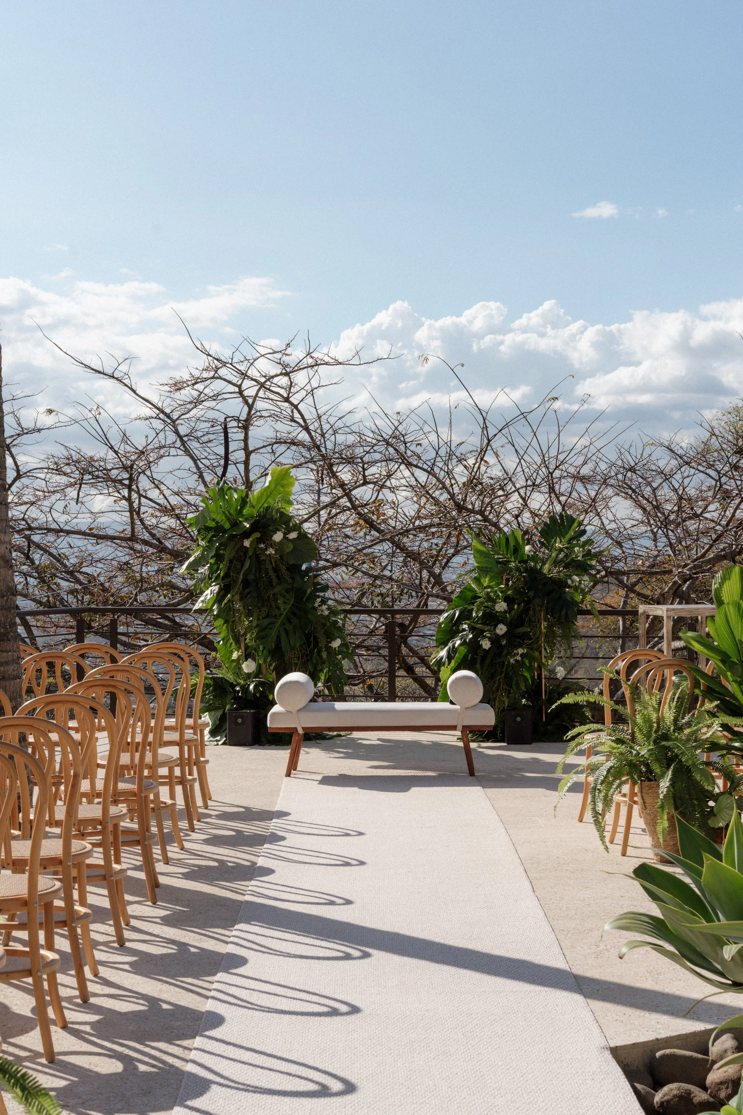 Espacio de ceremonia al aire libre con sillas de madera, pasillo blanco y decoración vegetal rodeada de naturaleza.