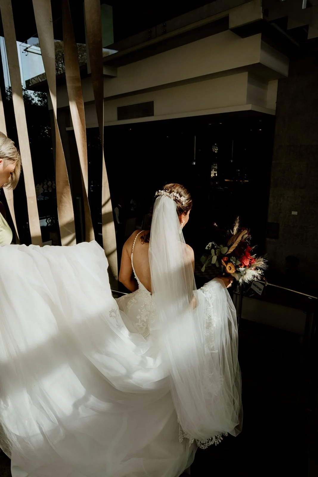 A bride in a white wedding dress with a veil, holding a bouquet, sitting indoors with sunlight casting shadows.