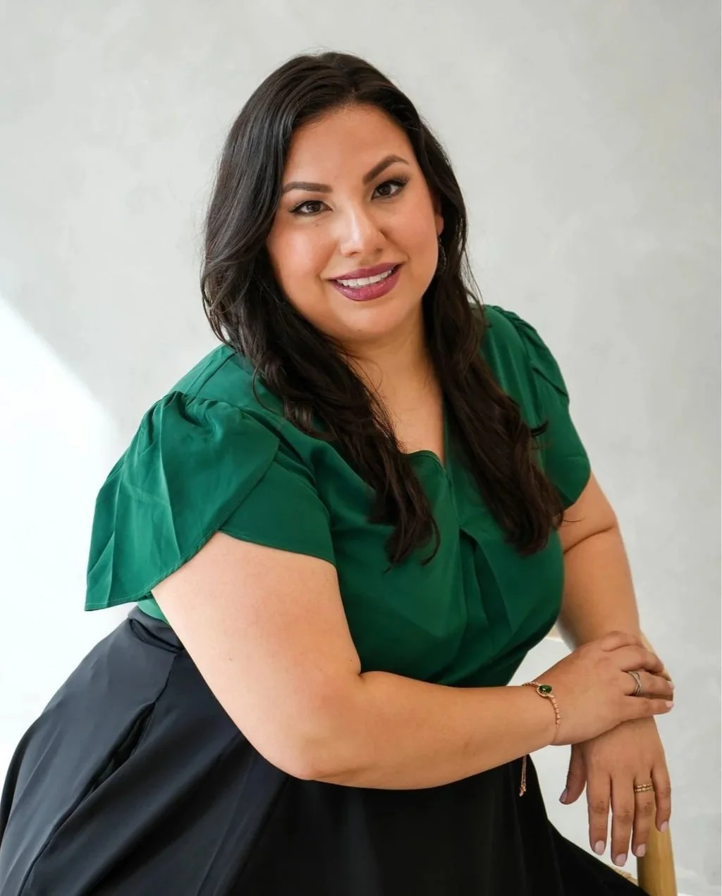 A woman with long dark hair smiling, wearing a green top and black skirt, seated against a light background.