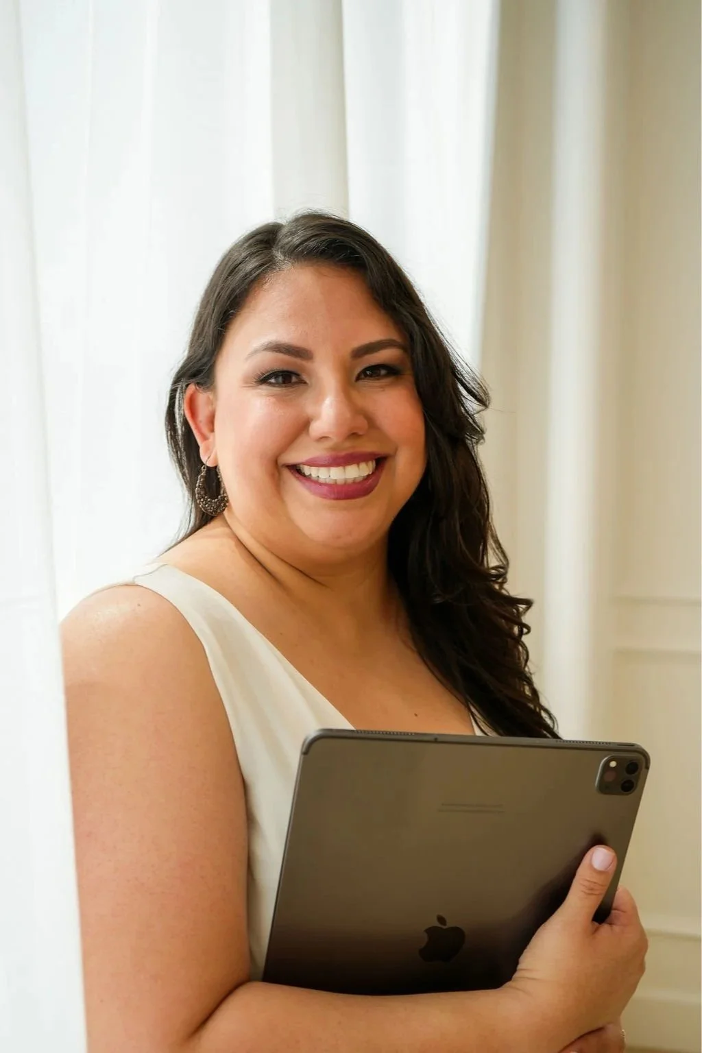 A woman smiling and holding an iPad, standing near white curtains.