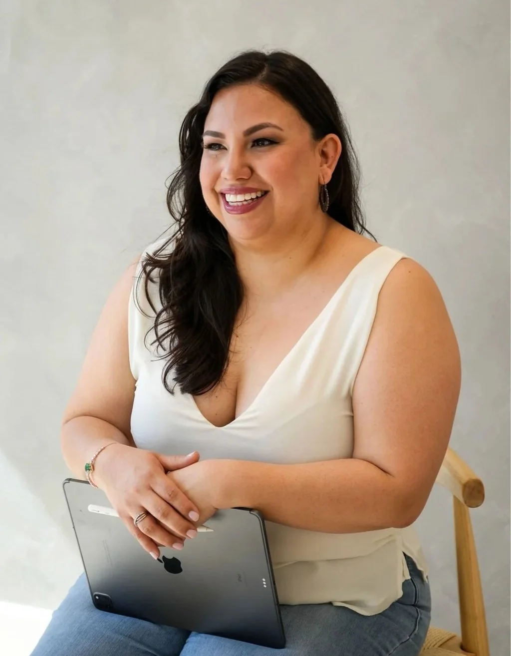 A woman with dark hair and earrings sitting on a wooden chair, holding an iPad and smiling, wearing a white top and jeans.