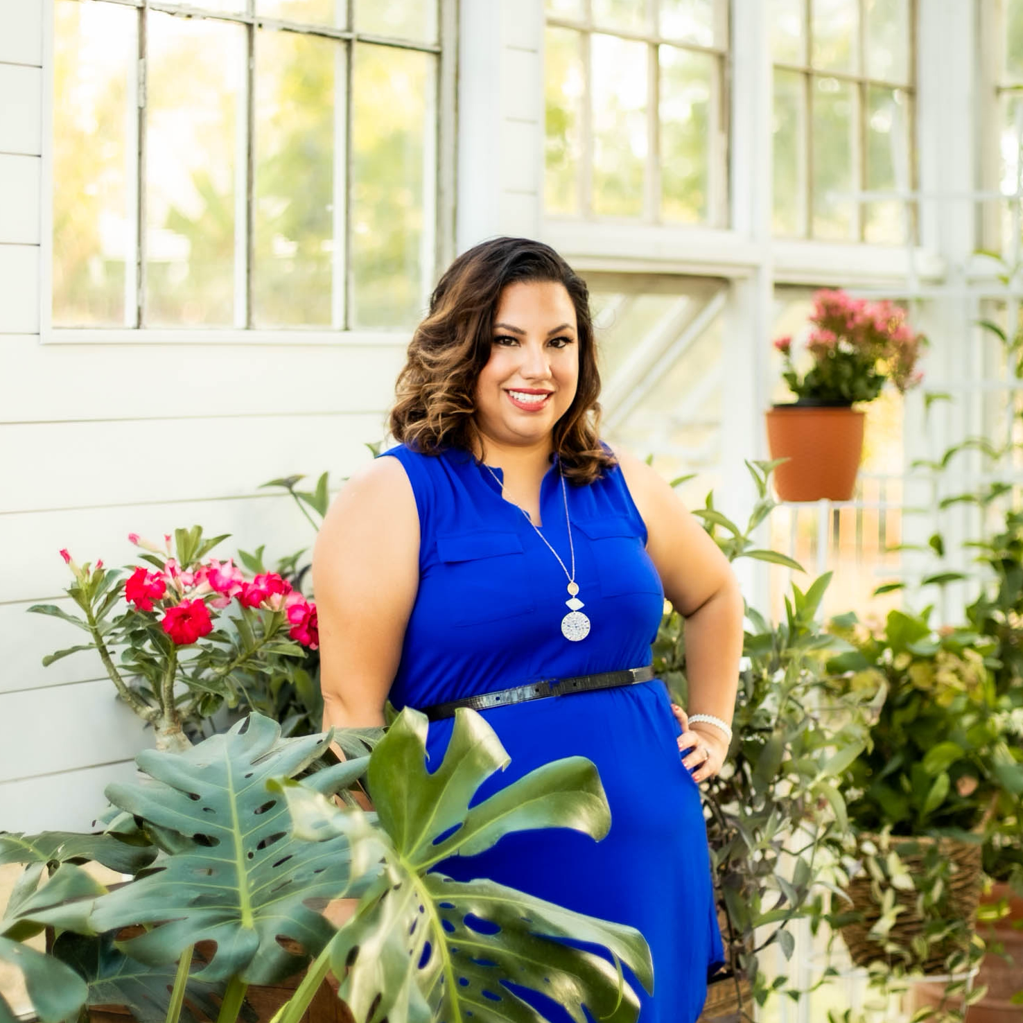 A woman in a blue dress standing in a greenhouse with green plants and pink flowers around her.