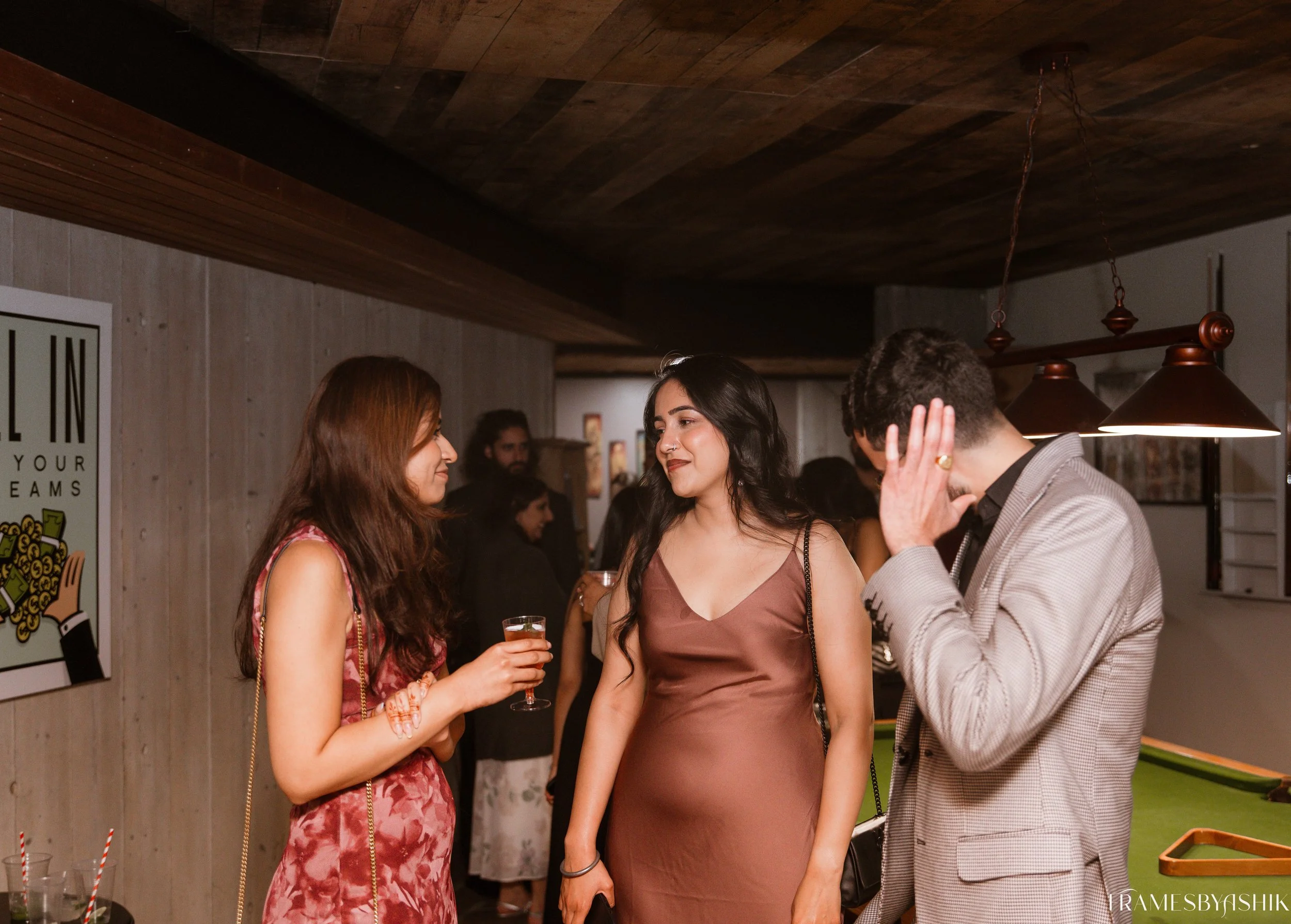 Three young adults, two women and a man, are socializing at a party, standing near a pool table, with drinks in hand, engaged in conversation.