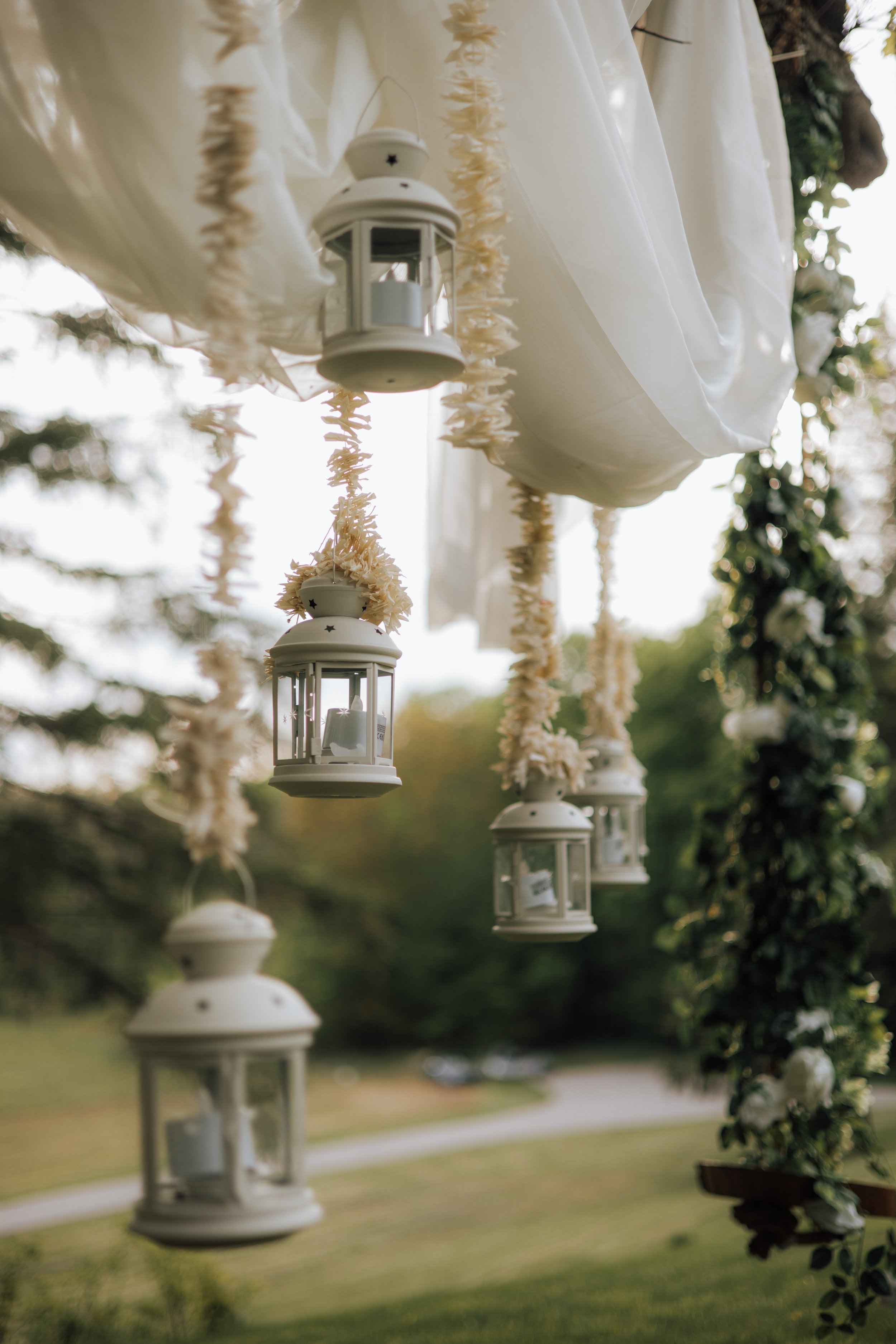 Hanging white lanterns with candles inside, decorated with cream-colored flower garlands, under white fabric canopy with blurred greenery background.