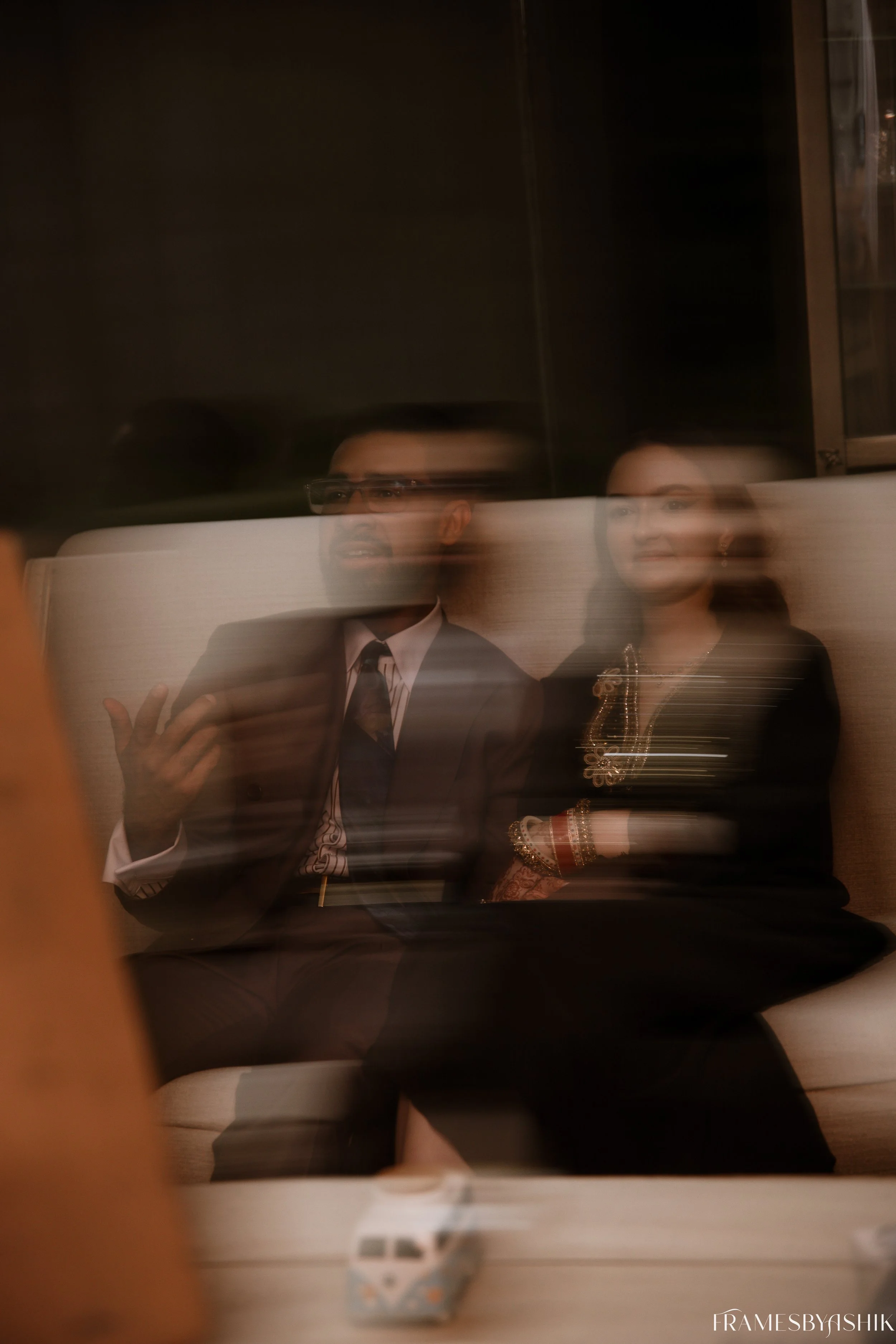 Two people sitting on a beige couch, viewed through a glass surface, creating a blurred double image. The man on the left is wearing glasses and a suit, gesturing with his right hand. The woman on the right is dressed in traditional clothing with jewelry, sitting with her arms crossed.