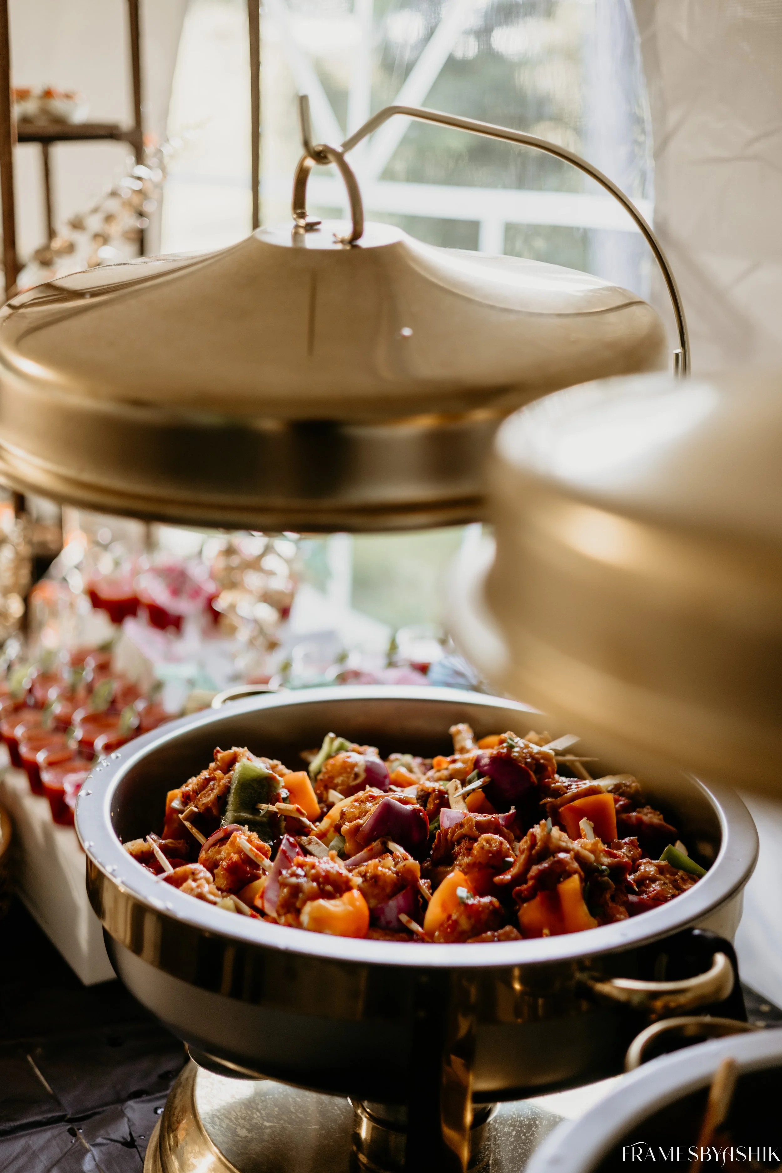 A buffet-style serving dish with cooked vegetables and meat, placed beneath a metallic heat lamp, in a bright room with large windows.