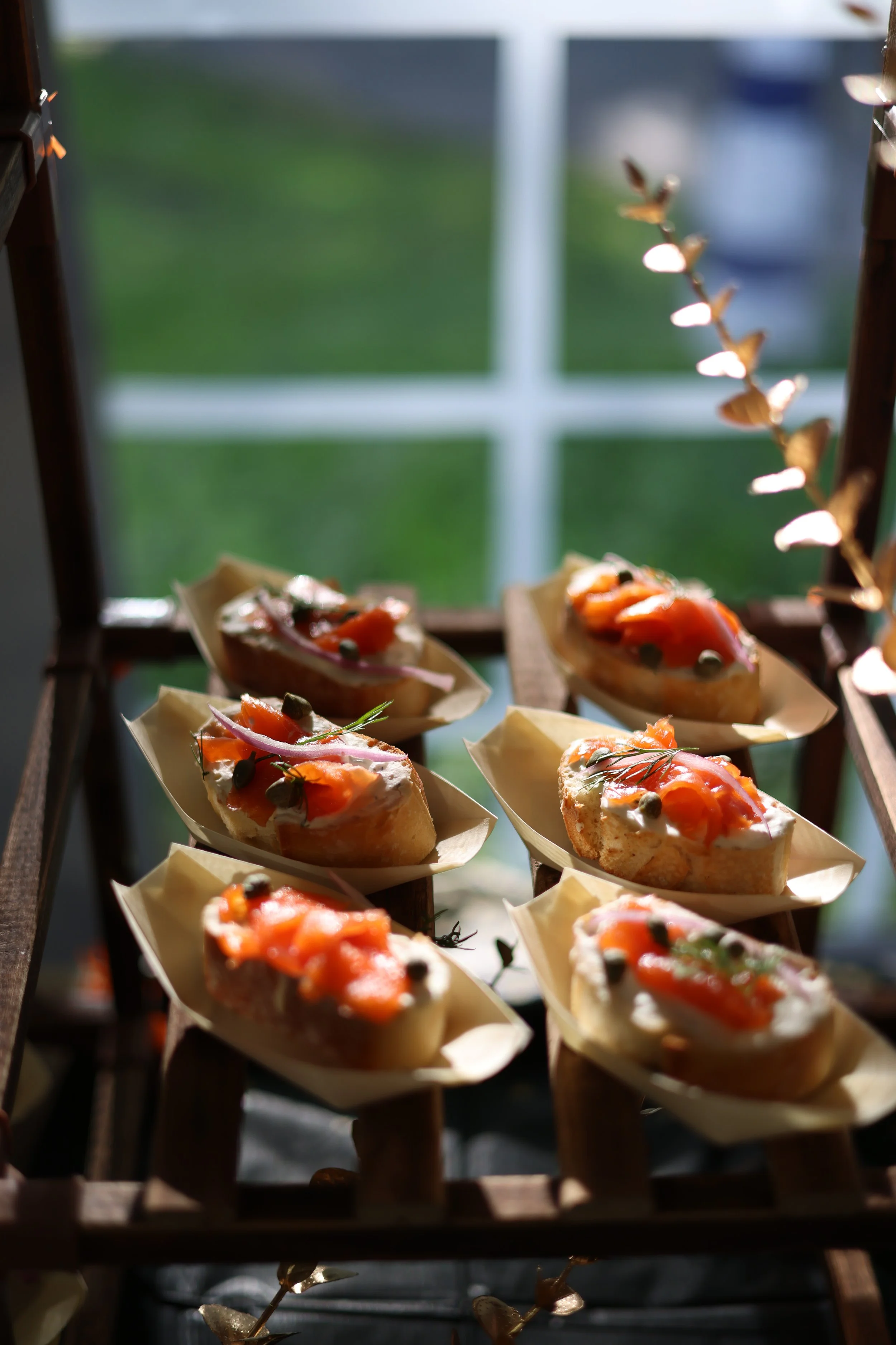 Close-up of sliced bread topped with smoked salmon, capers, red onions, and herbs, arranged on a wooden rack with a blurred window in the background.