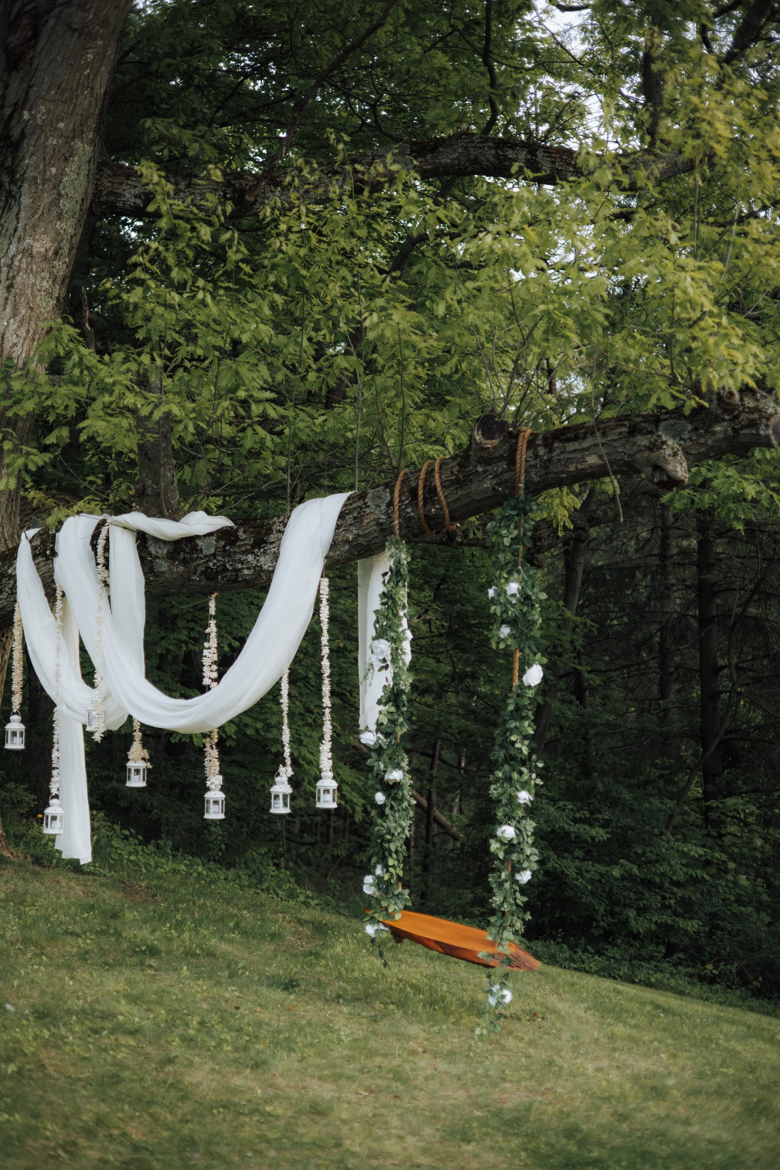 A decorative outdoor setup featuring a large tree with fabric, lanterns, and floral arrangements hanging from a branch, including a swing with flowers, greenery, and an orange cushion, set in a wooded area.