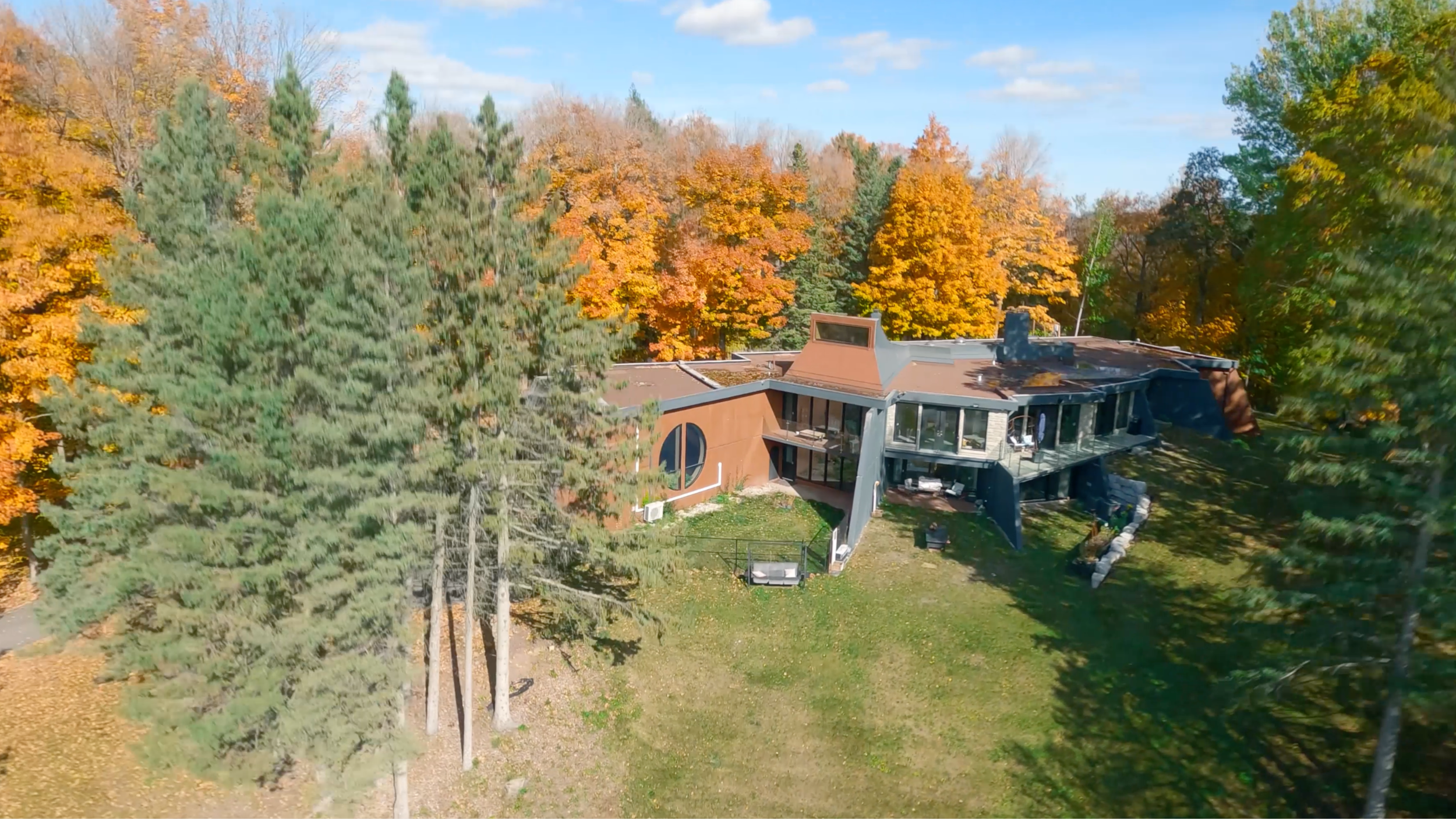 Aerial view of a modern house surrounded by colorful autumn trees with orange, yellow, and green leaves, and a lawn in the foreground.