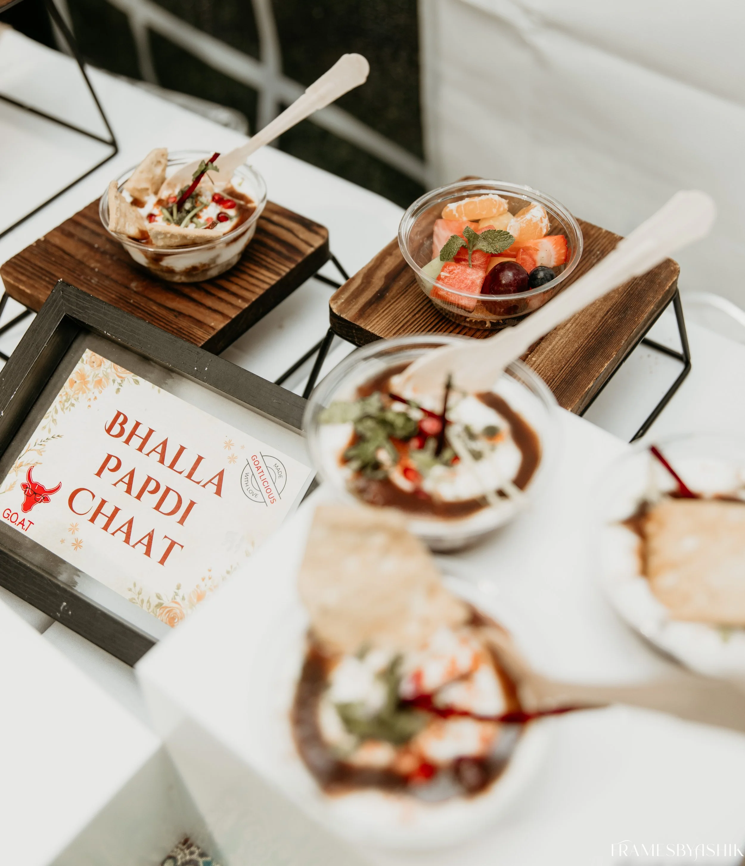 Bowls of Indian desserts with fruit and garnishes, displayed on a white table with a sign reading "Bhalla Paddi Chaat."