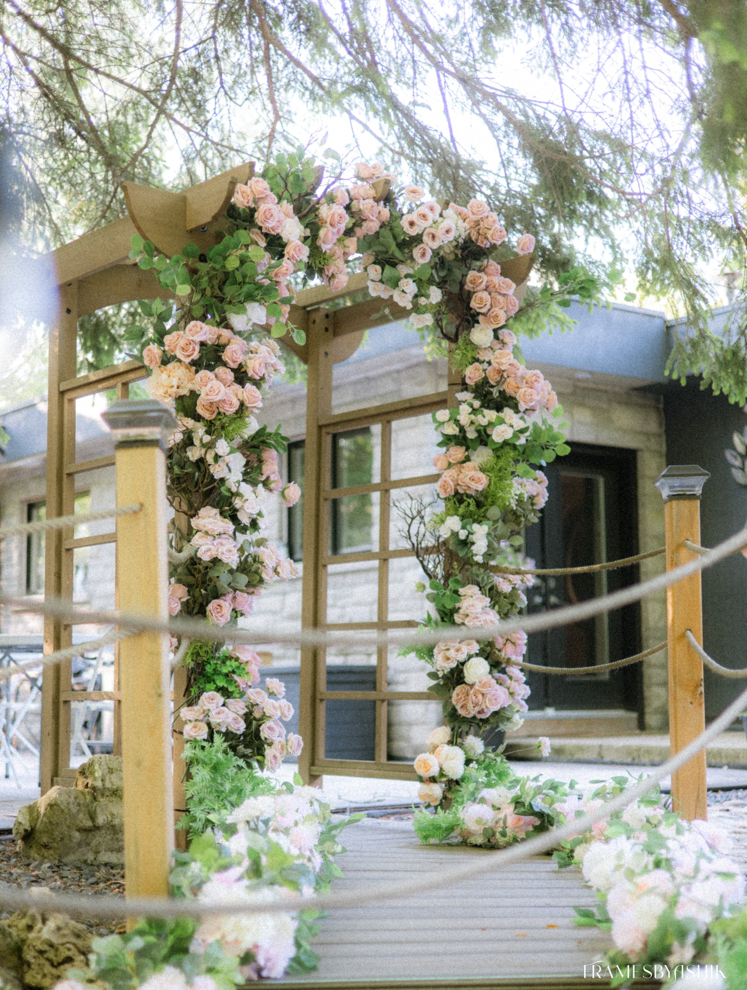 Flower arch with pink and white roses, greenery, and wooden structure on a small bridge, outdoors with trees and building in the background.