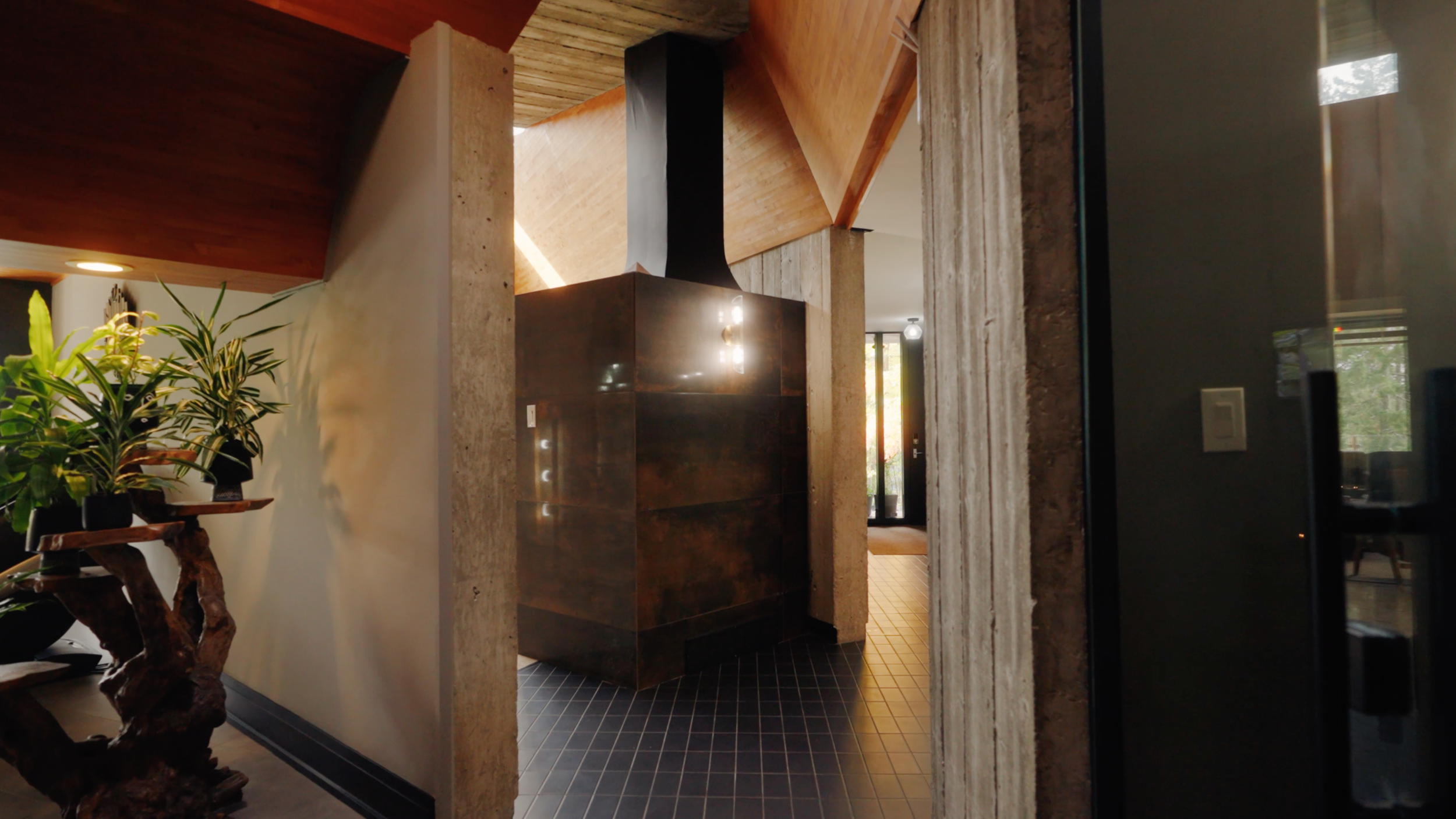 Interior view of a modern house with a wooden ceiling, a large potted plant on a wooden stand, and a dark-colored kitchen island or cabinet near a hallway with a black tiled floor and a glass door.