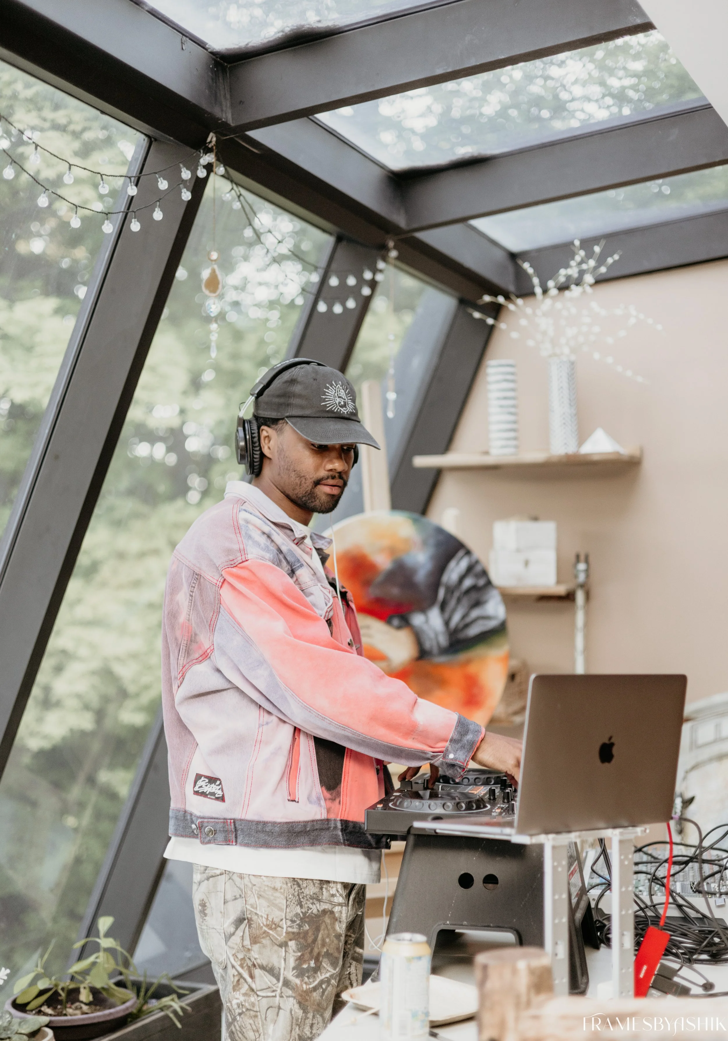 A man wearing a cap and headphones standing at a DJ setup with a MacBook laptop, inside a glass greenhouse or sunroom with plants, shelves, and decorations.