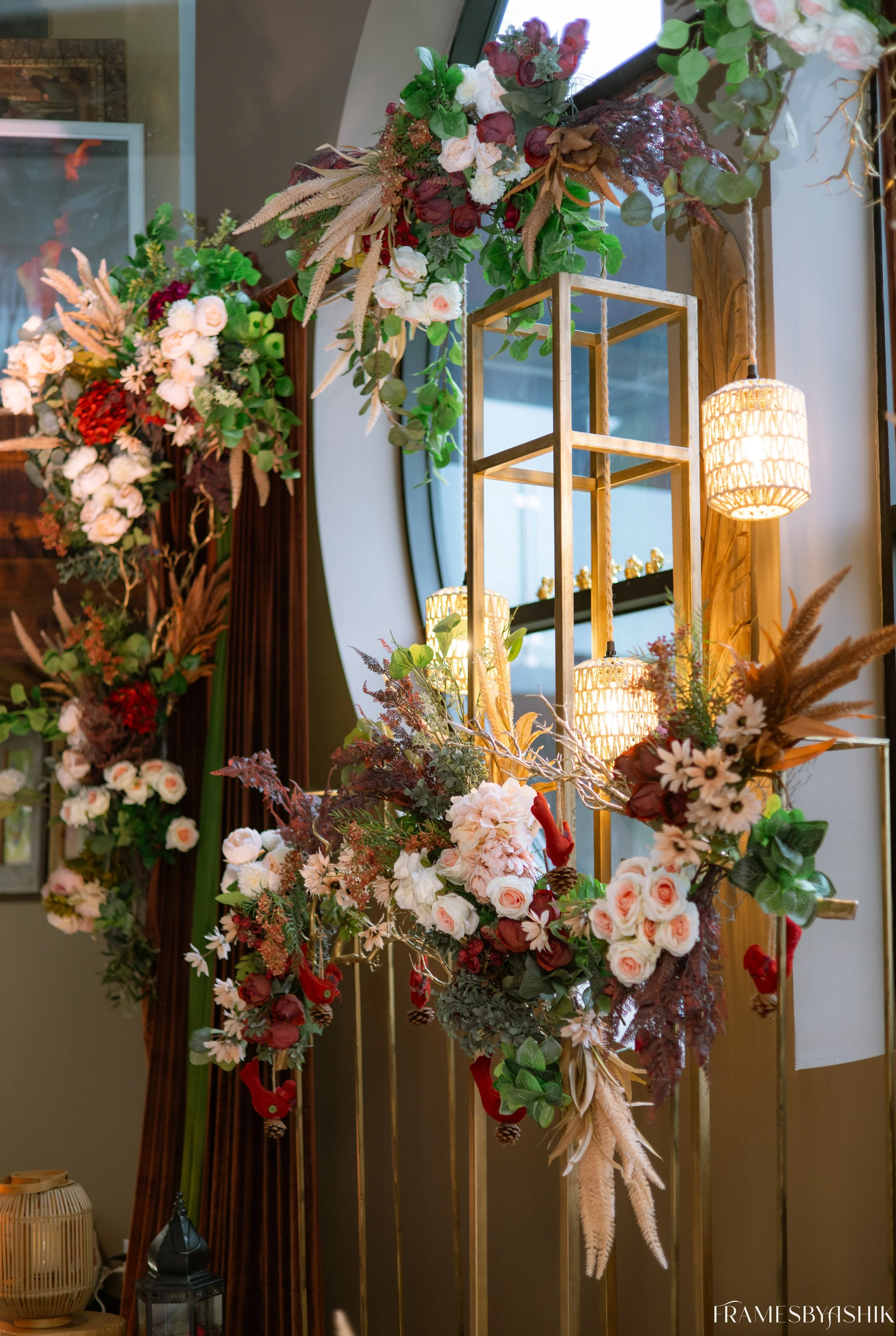 Decorative floral arrangement with pink, white, and red flowers, greenery, and dried elements, adorned around a gold metallic frame with hanging wicker-style lamps, near a mirror and window.