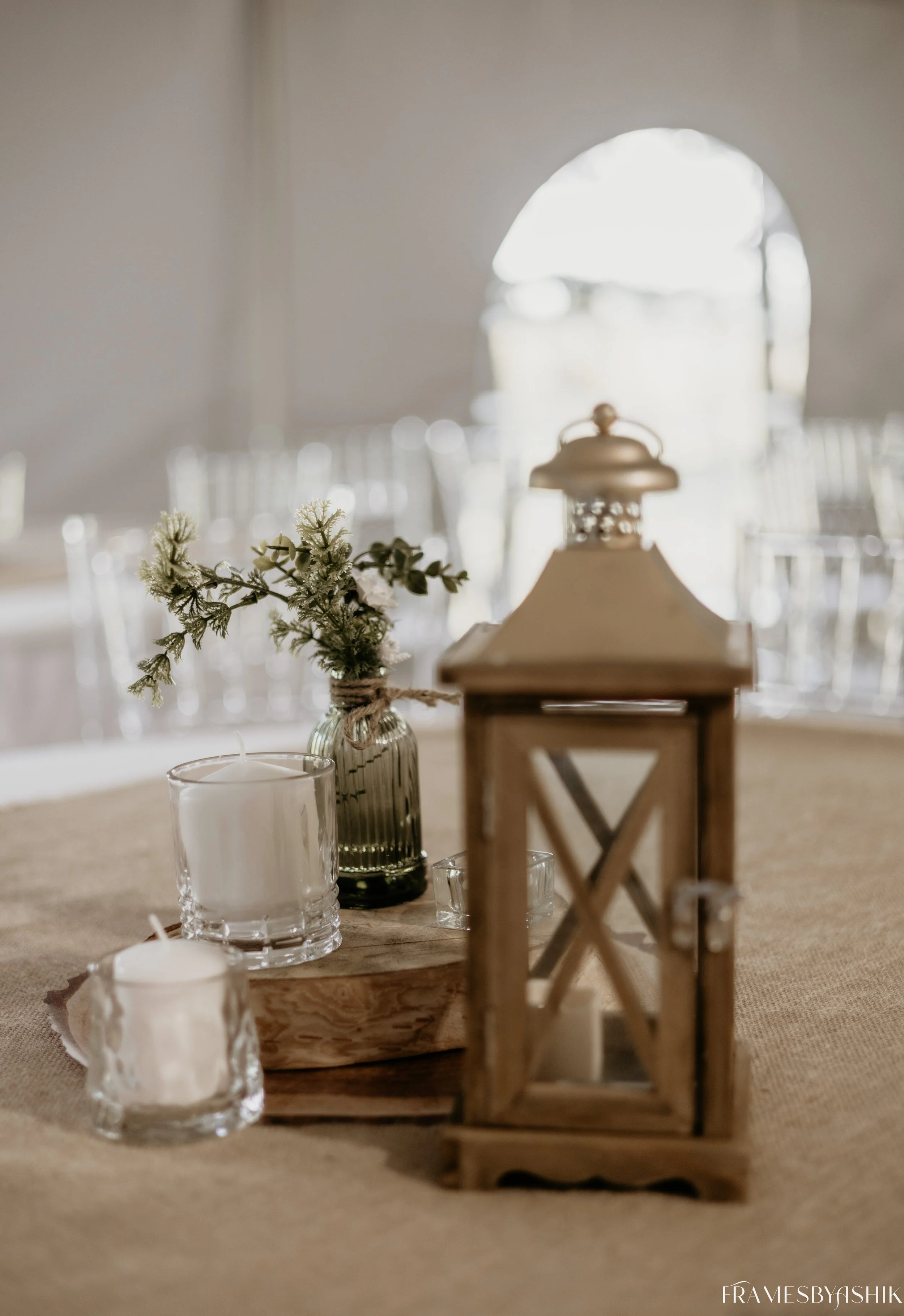 A table centerpiece featuring a wooden tray with candles in glass holders, a small green vase with a sprig of greenery, and a rustic lantern on a beige tablecloth, with a blurred background of chairs and a window.