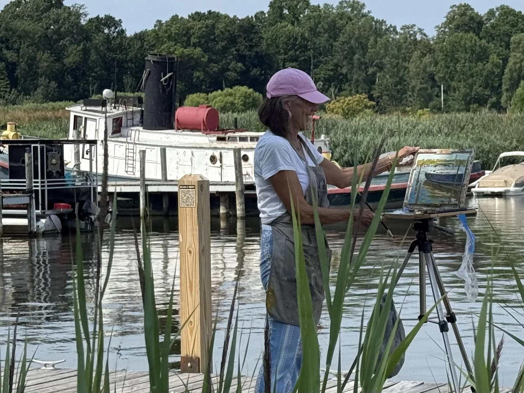 A woman wearing a purple cap and casual clothes is painting a large watercolor of a boat on an easel, standing near a body of water with boats and a dock. Tall grass in the foreground partially obscures the scene. Trees line the background.