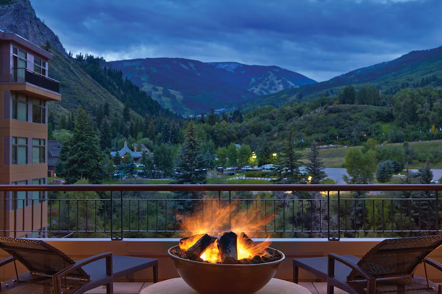 A balcony with a fire pit and two chairs overlooking a scenic mountain landscape with buildings, trees, and a lake.
