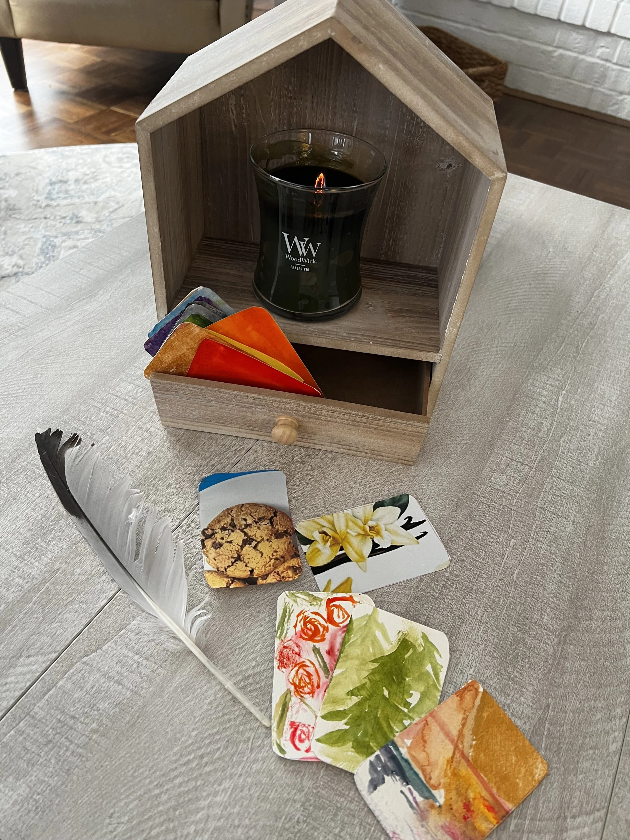 A small wooden cabinet with a candle inside and a drawer containing colorful paper or cards with floral and leaf patterns, set on a light-colored wooden table.