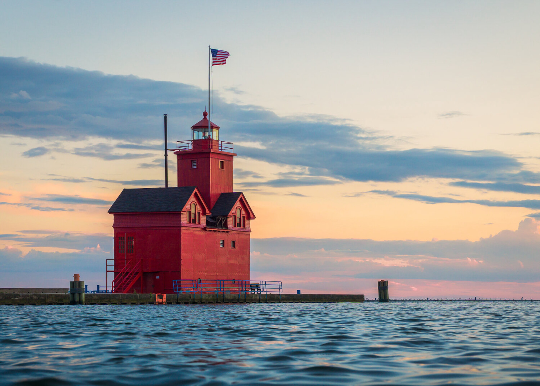 Red lighthouse on a pier with an American flag flying at sunset over the water and a cloudy sky.