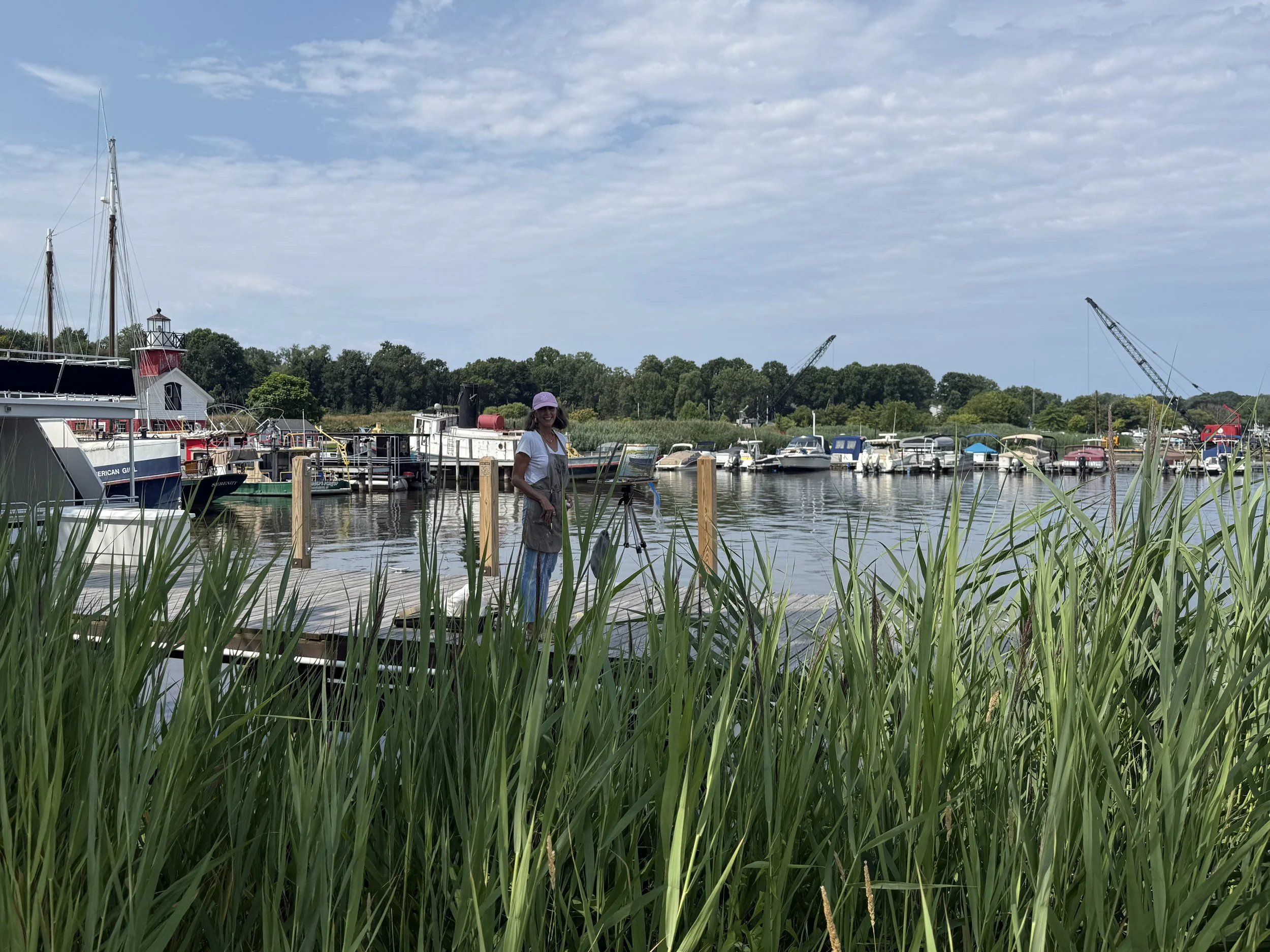 A person standing on a dock near a marina with boats and sailboats, surrounded by green grass and trees under a partly cloudy sky.