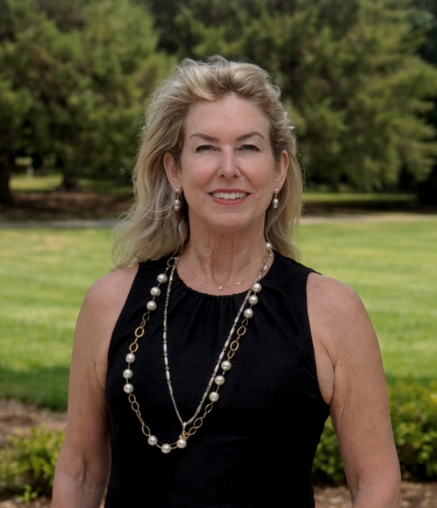 A middle-aged woman with blonde hair wearing a black sleeveless dress and layered pearl and gold necklaces, standing outdoors in a park with green trees and grass in the background.
