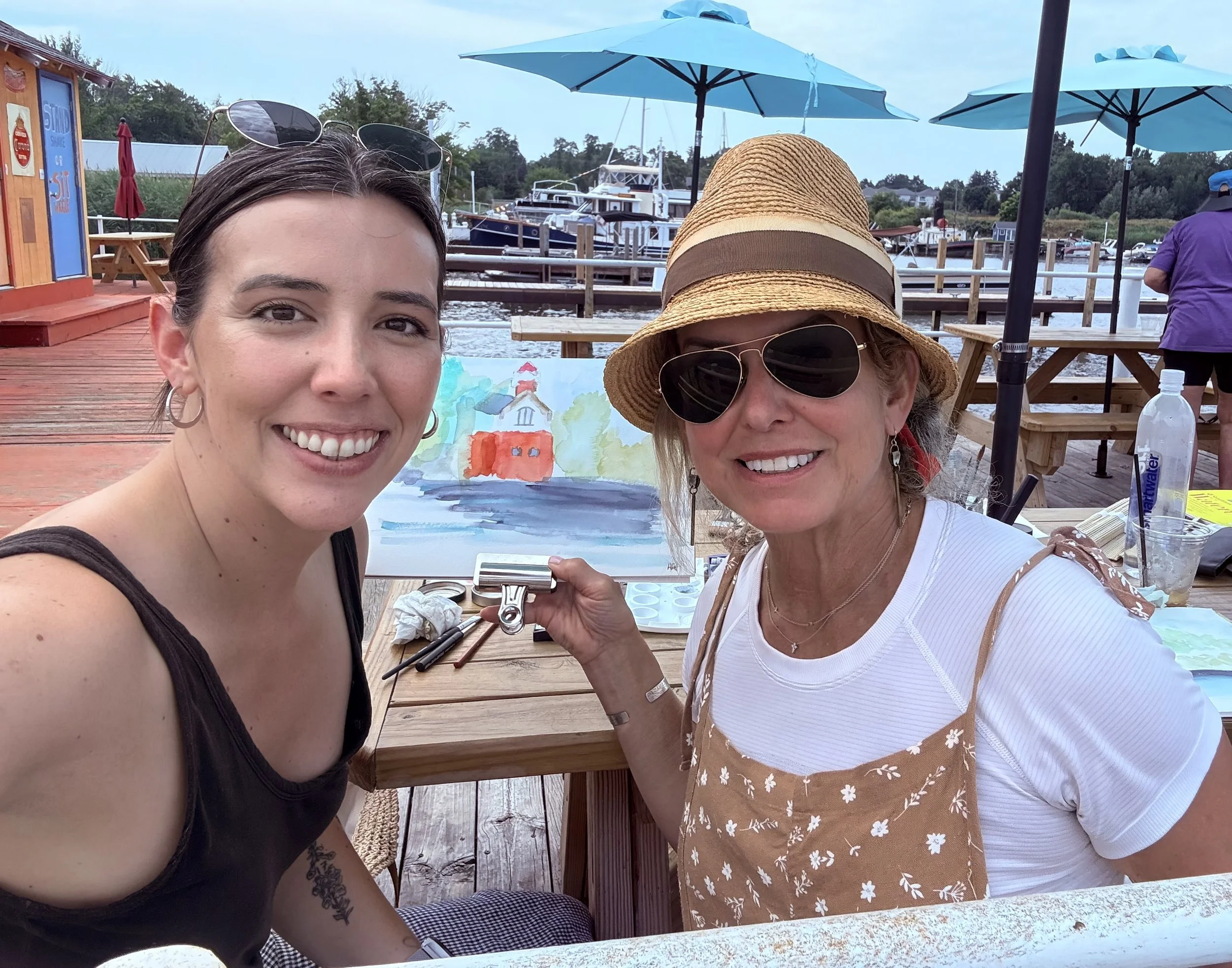 Two women smiling at a marina, with one holding an art piece of a colorful lighthouse painting, surrounded by wooden tables and blue umbrellas.
