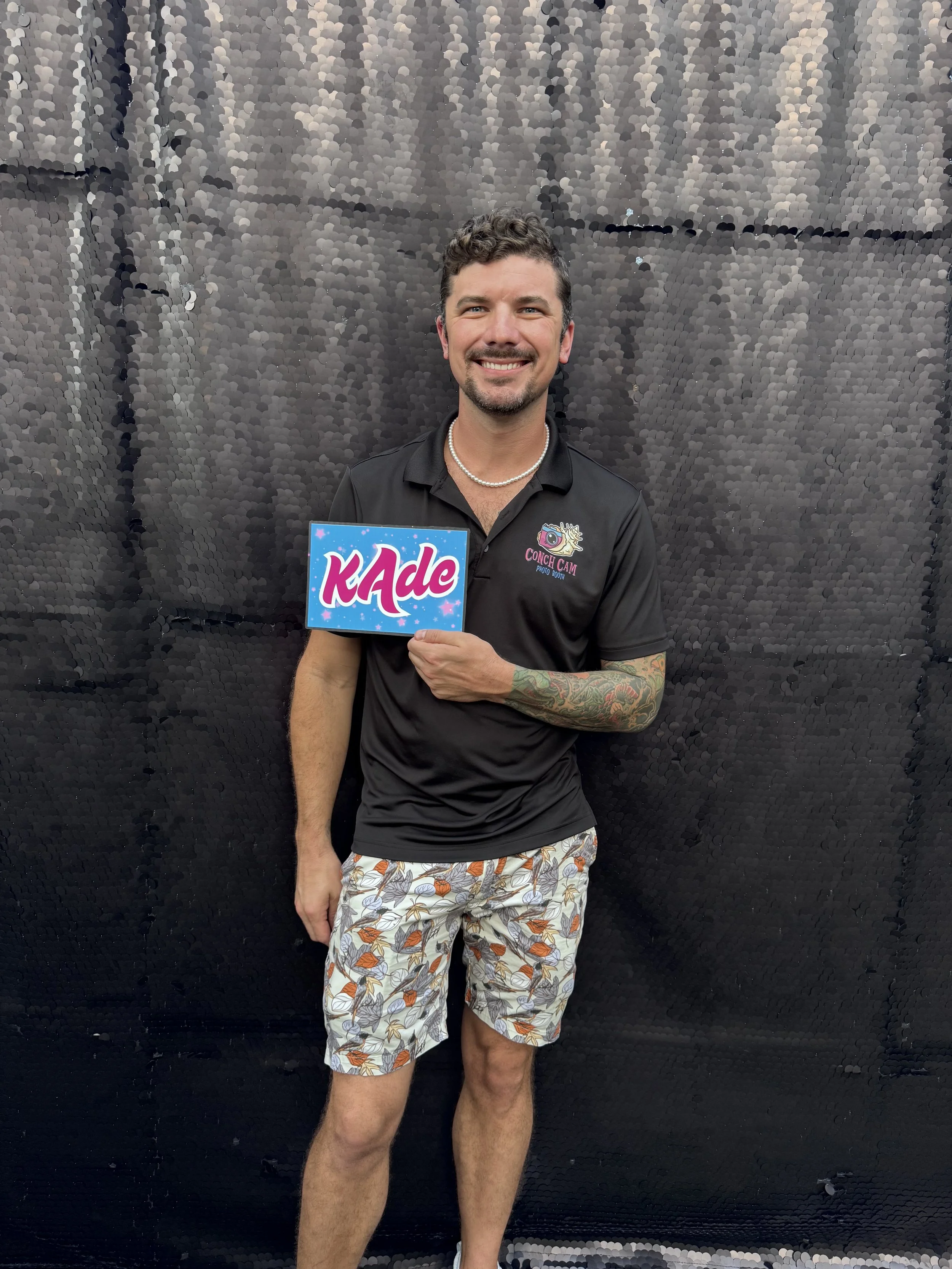 A smiling man holding a sign with the word 'KADE' in colorful letters, standing in front of a black textured wall. He wears a black polo shirt, patterned shorts, a pearl necklace, and has tattoos on his left arm.