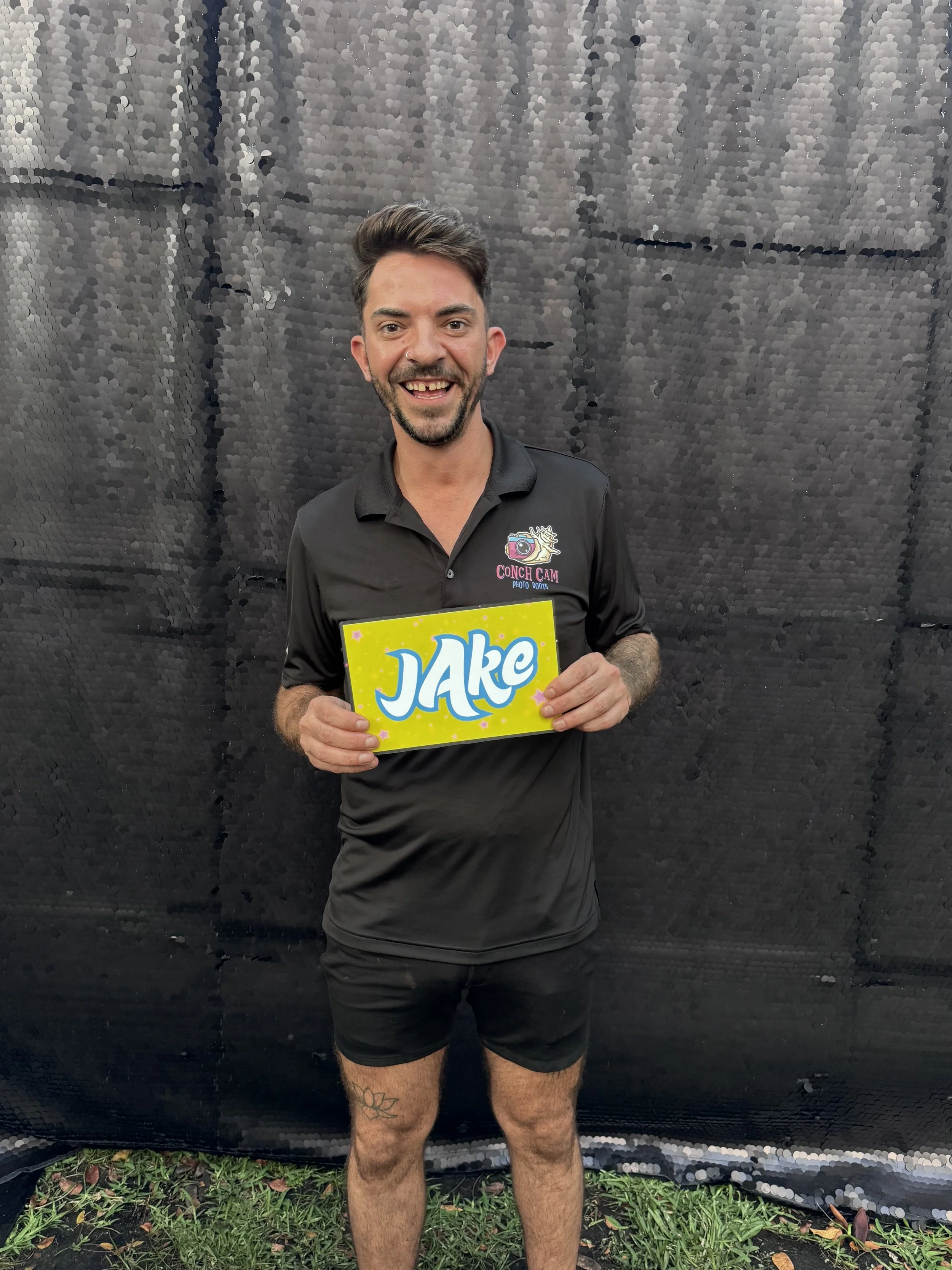 A smiling man in a black polo shirt and shorts holding a bright yellow sign with the word 'Jake' in blue, standing outdoors against a black backdrop.