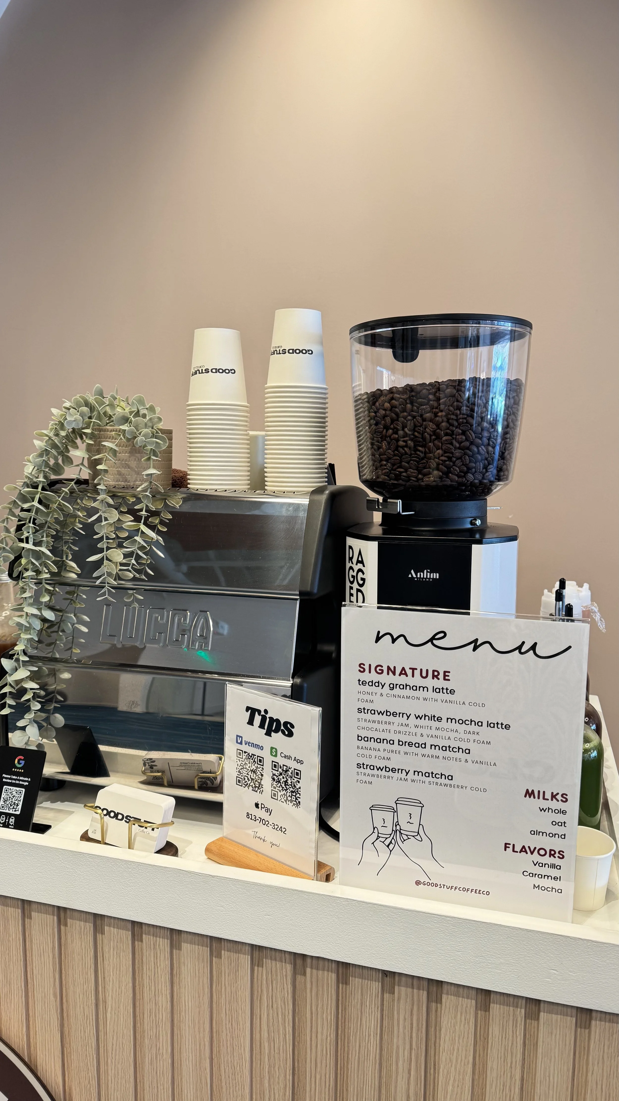 A coffee shop counter with a LUCIA espresso machine, a jar filled with coffee beans, disposable cups, a small plant, and a menu listing various coffee drinks and flavors.