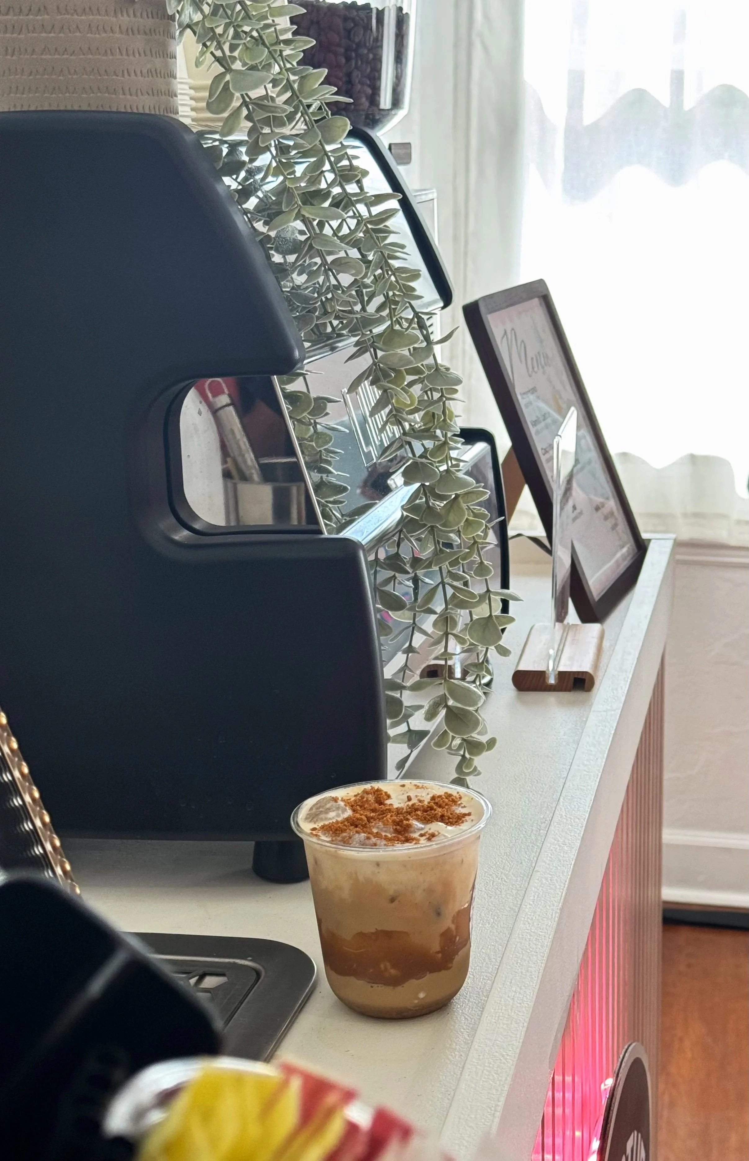 A whipped coffee drink topped with cinnamon or brown sugar in a clear plastic cup, sitting on a white counter in a cafe or coffee shop.