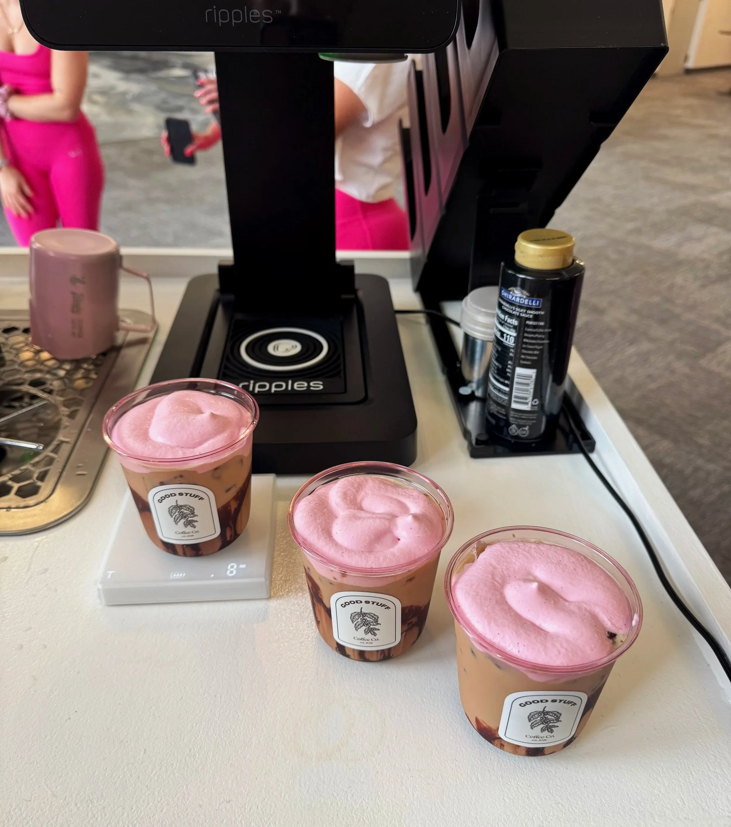 Four beverages with pink foam on top, placed on a white surface. There are three identical cups with a label reading 'Good Stuff Coffee Co.' and what appears to be a chocolate drizzle. A digital scale is beneath one of the cups. In the background, th